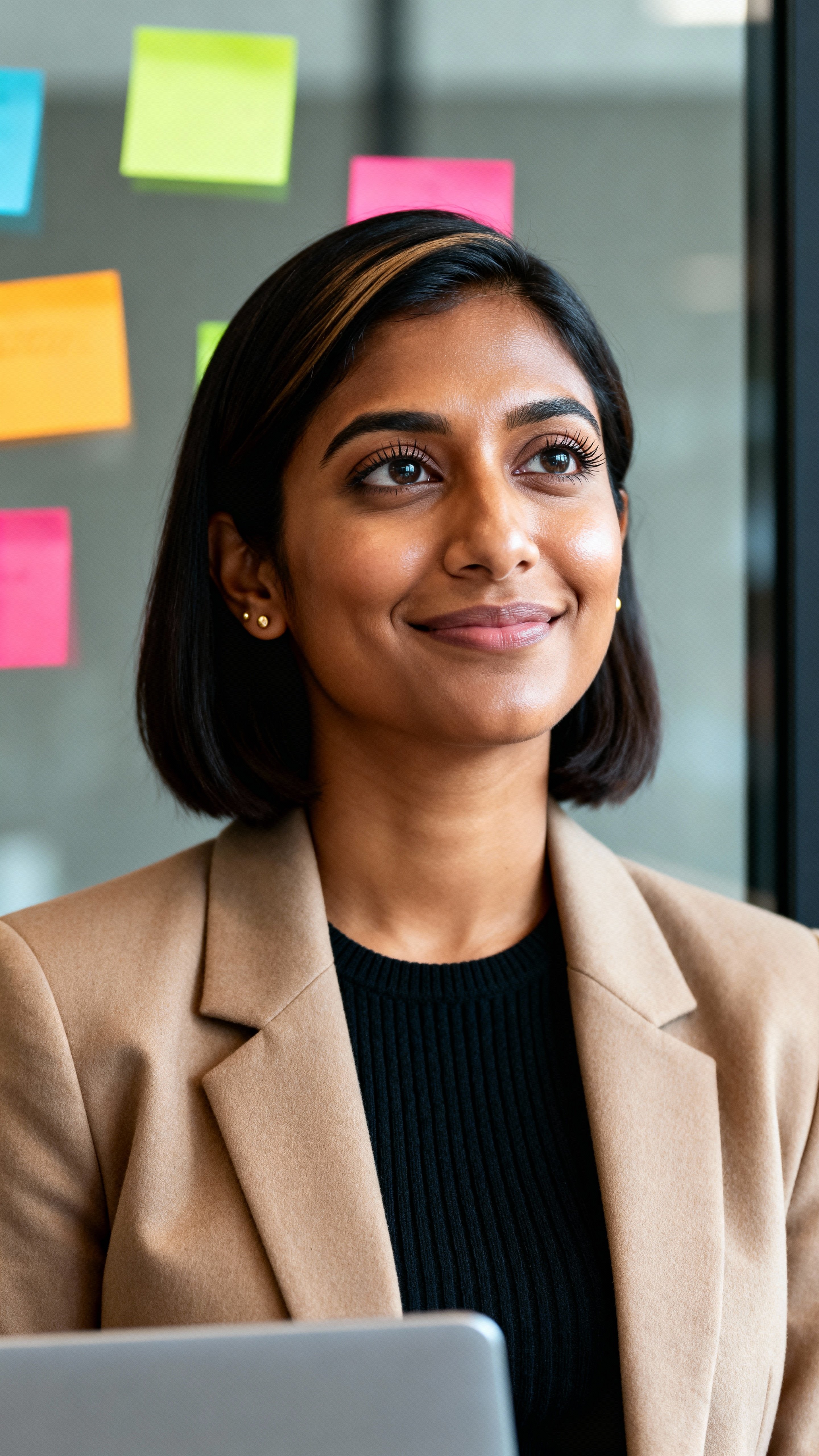 Aparna, a South Asian American product manager in her early 30s, smiles mid-thought by a glass wall in a Boston coworking space wearing an oatmeal blazer and black knit, warm brown skin with minimal gold studs, crisp iris detail and natural