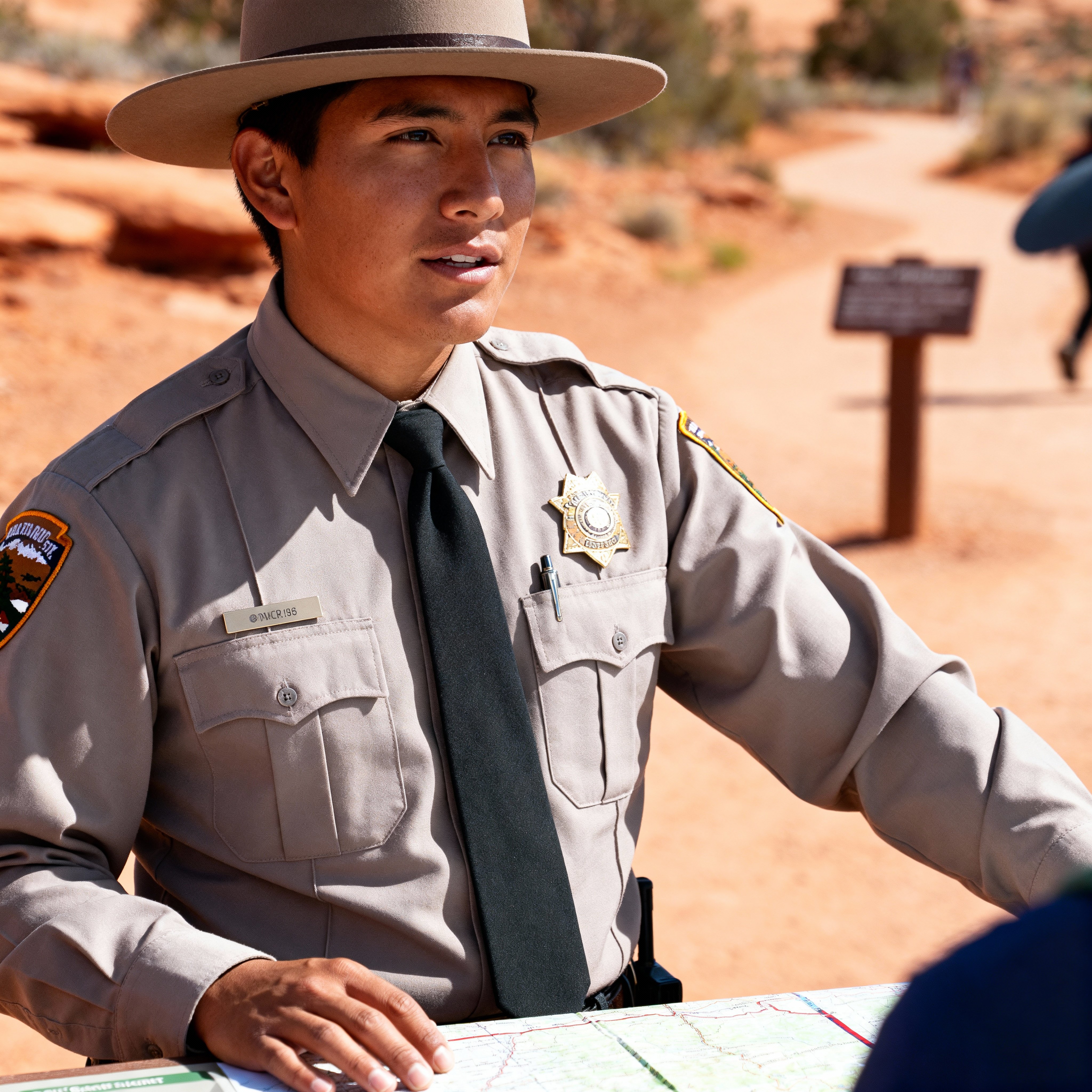 Ashkii Begay, half-body in a pressed uniform and flat-brim hat, rests a hand on a trail map and guides visitors with steady authority in clear light; sandstone (#c97b4a), rust (#d94f2b), cream (#f2e8d5), forest-green (#35524a), earth-brown