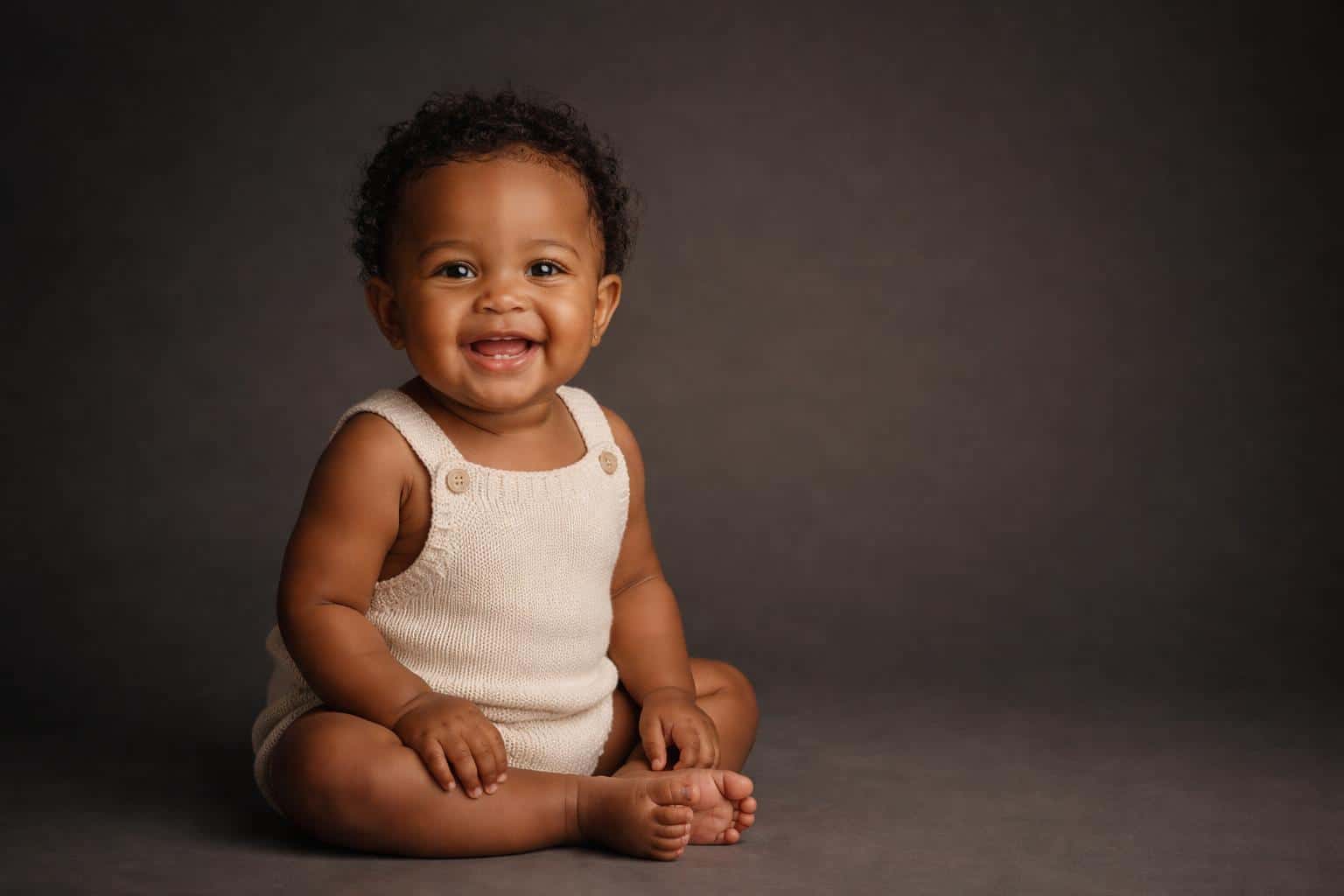 Wide 3:2 cinematic editorial studio portrait of a one-year-old baby girl with warm brown skin and short tight curls seated on a deep charcoal seamless backdrop.