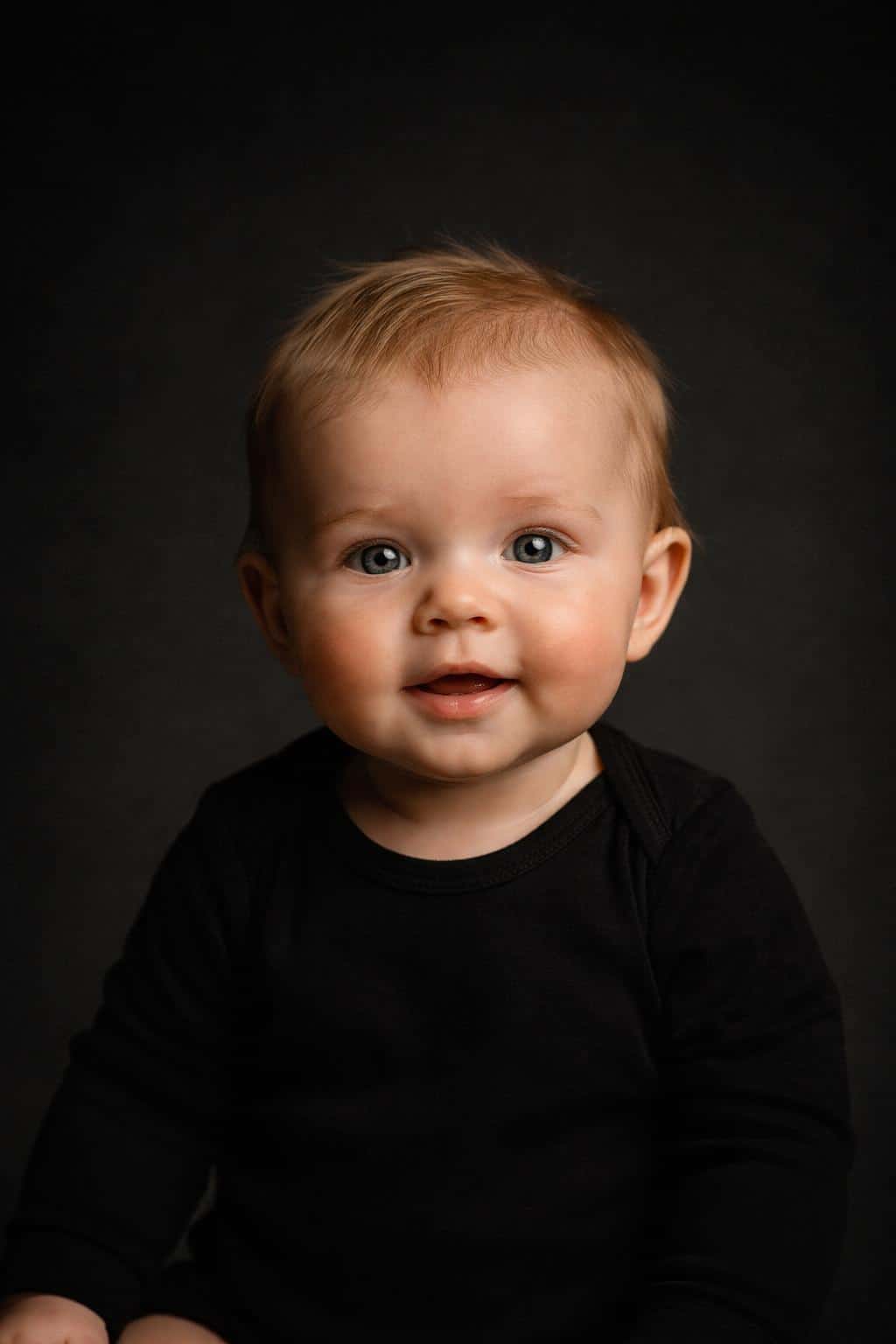 Cinematic 2:3 studio portrait of a six-month-old baby girl with freckled fair skin and wispy light hair, upright against a seamless dark gradient with warm amber highlights and cool charcoal shadows.