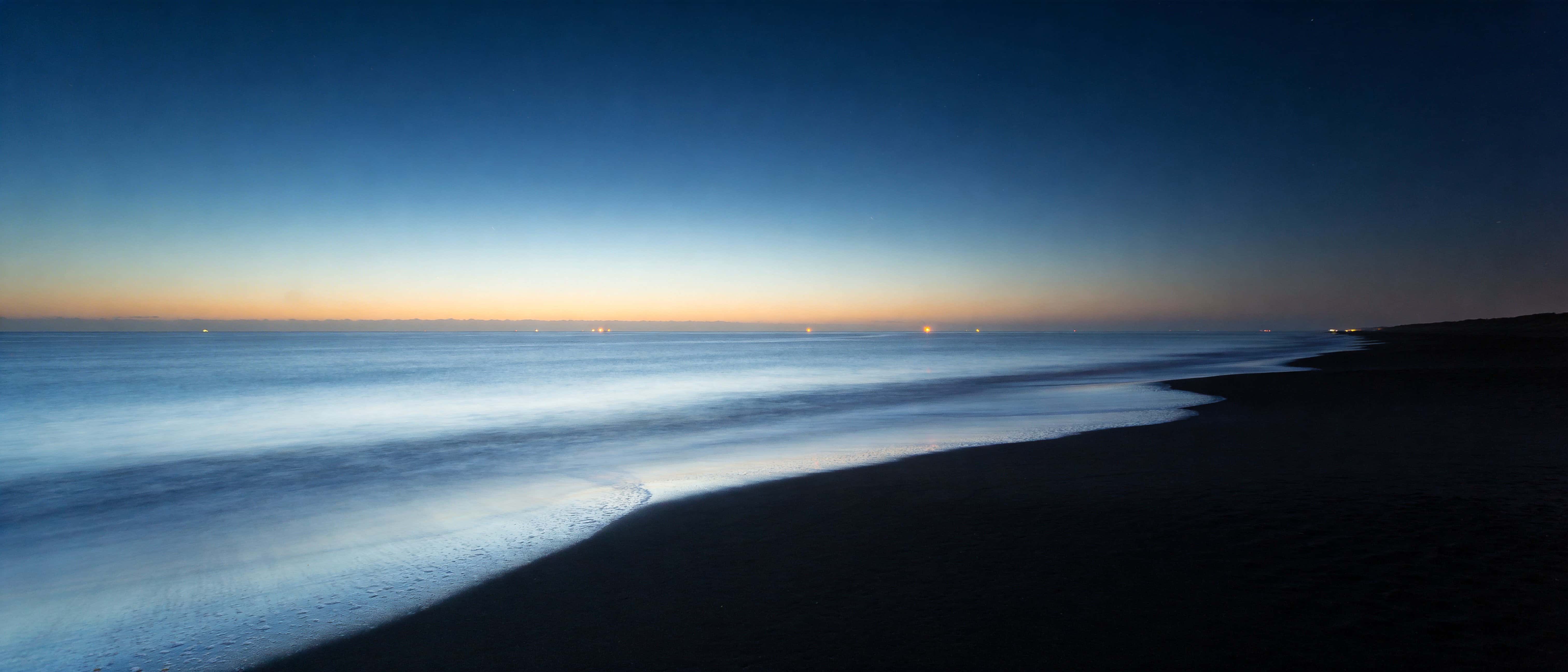 A 21:9 ultra-widescreen blue-hour beach photograph with a long sweeping shoreline, expansive negative space, calm steel-blue water, soft haze, and warm distant bokeh.