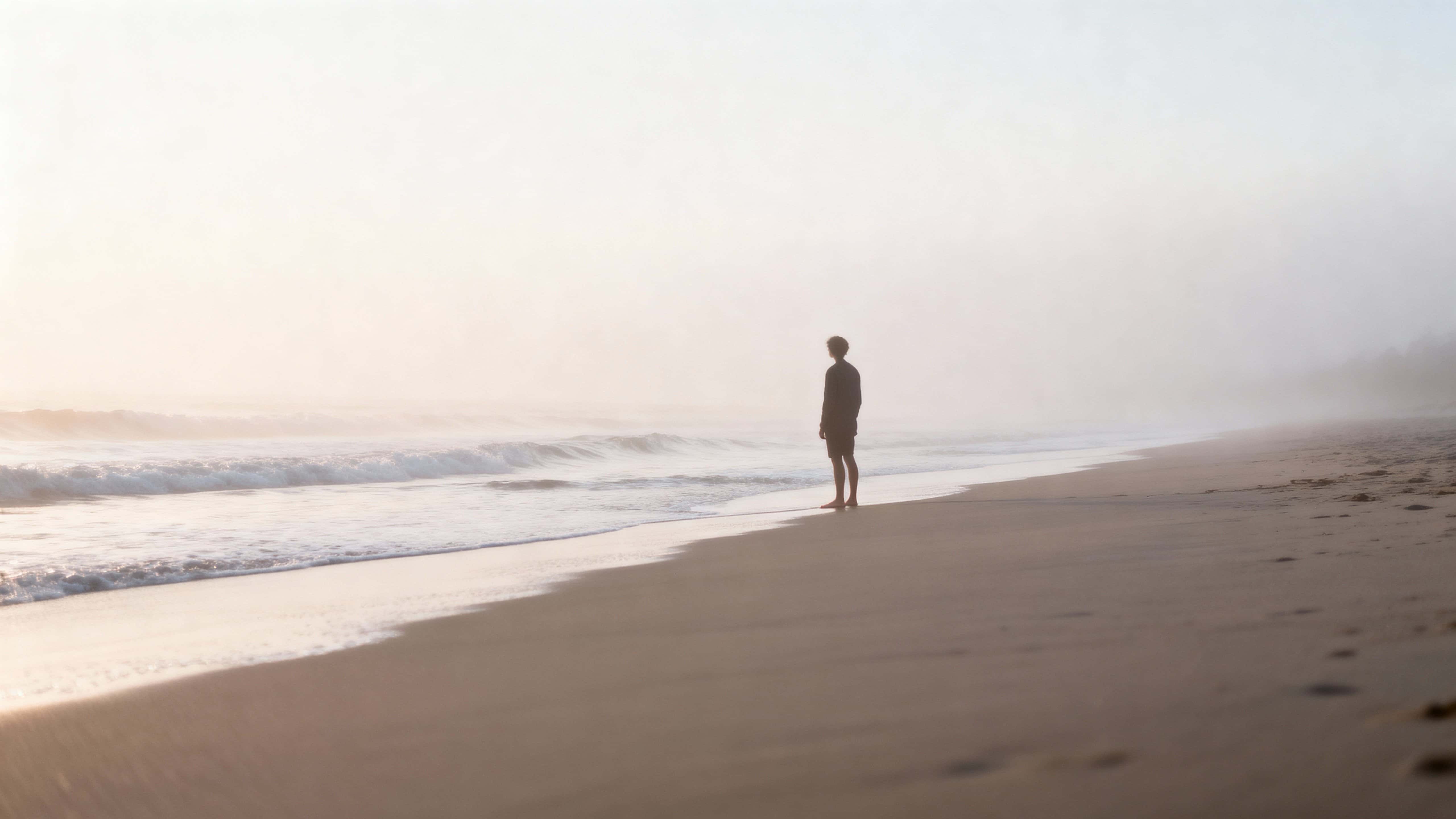 A lone figure stands barefoot at a calm, empty beach shrouded in soft morning fog with gentle waves kissing the shore and pastel tones rendered in realistic photographic detail.