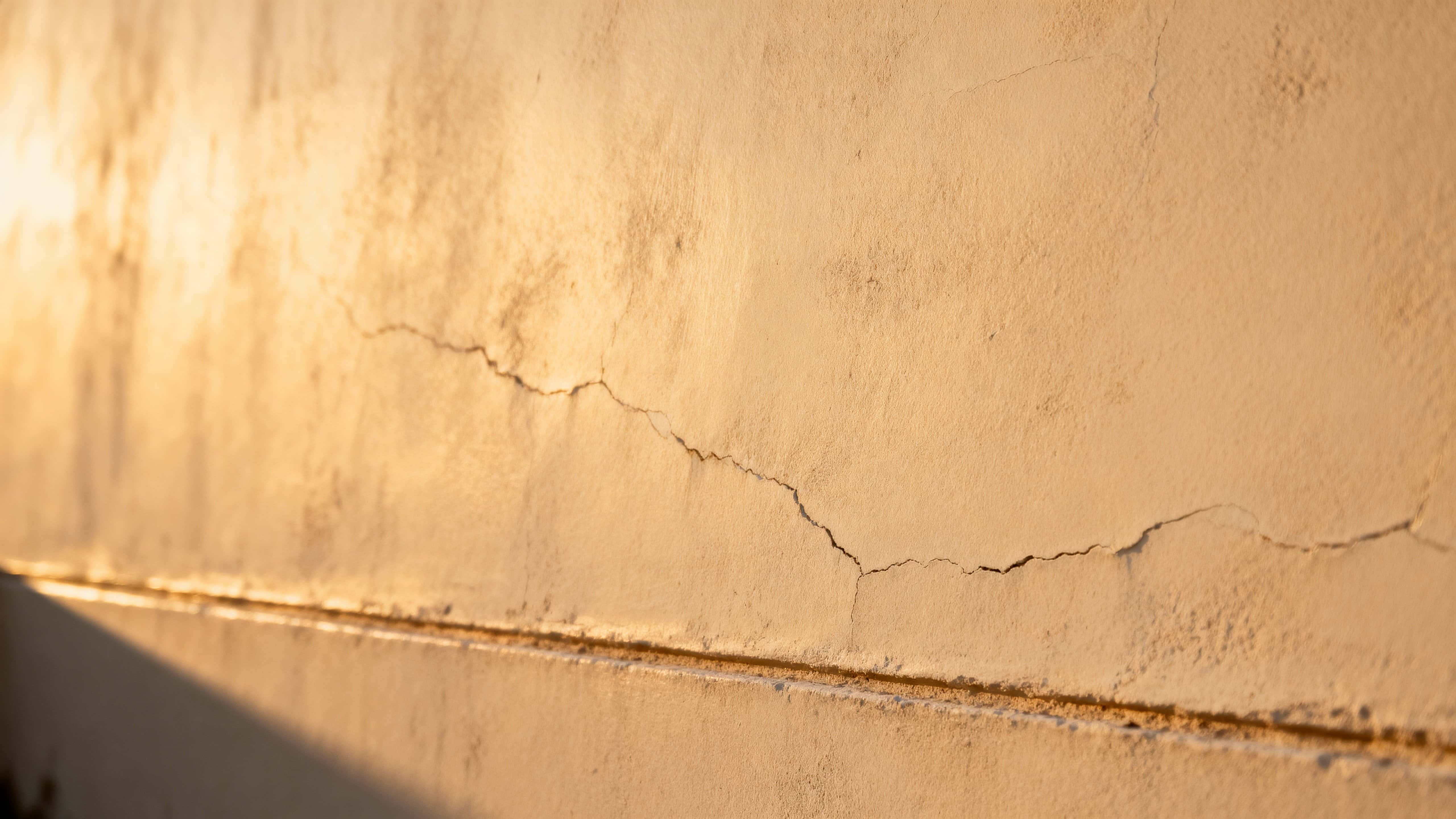 A soft beige wall surface is shown with warm sunlight sculpting subtle texture and shadow across a 16:9 frame, creating a calm, tactile study suitable for background or material reference photography.