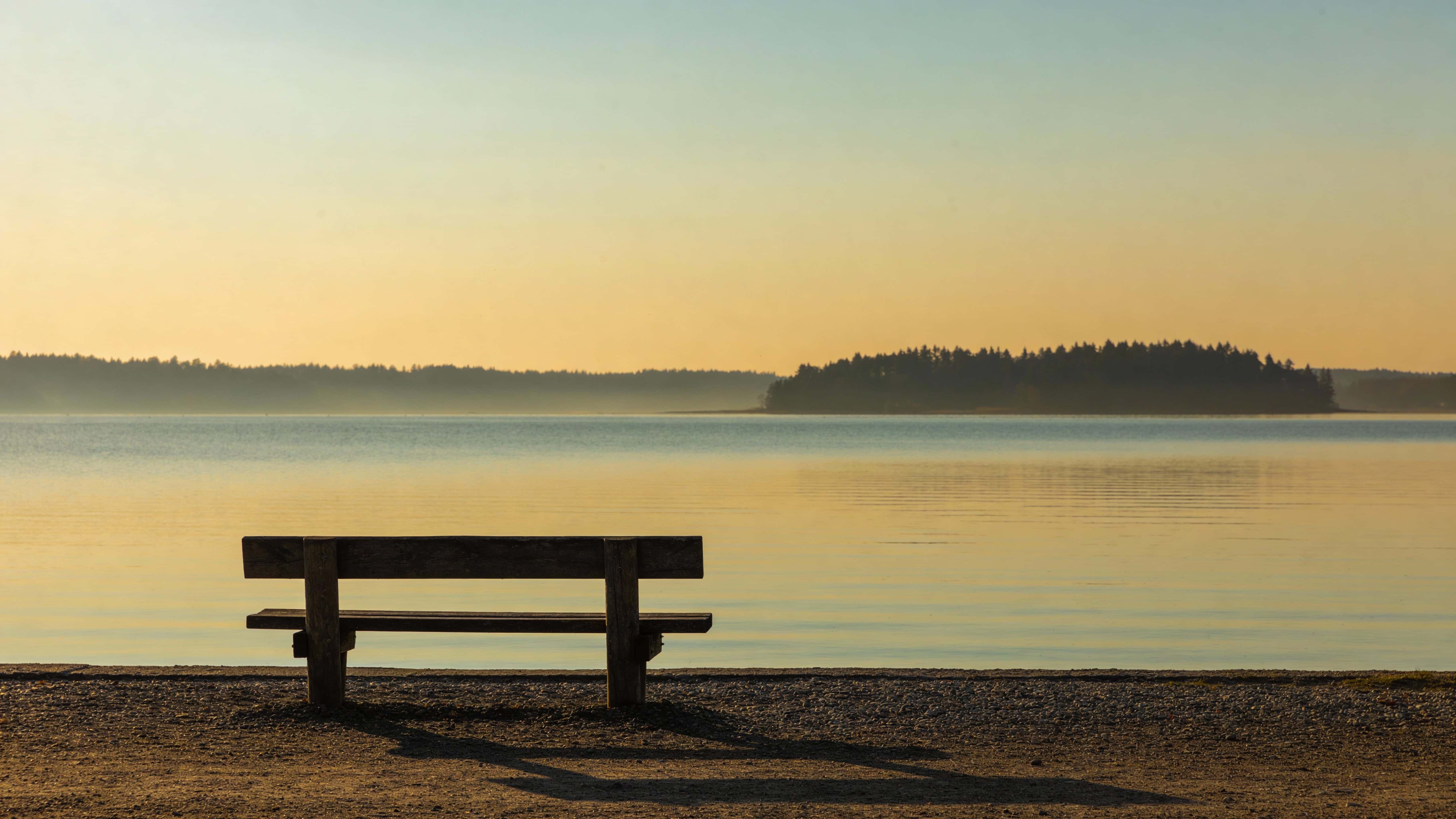 Solitary wooden bench beside a calm lake captured as a 16:9 landscape photograph with soft golden-hour light and clean composition for wallpaper use.