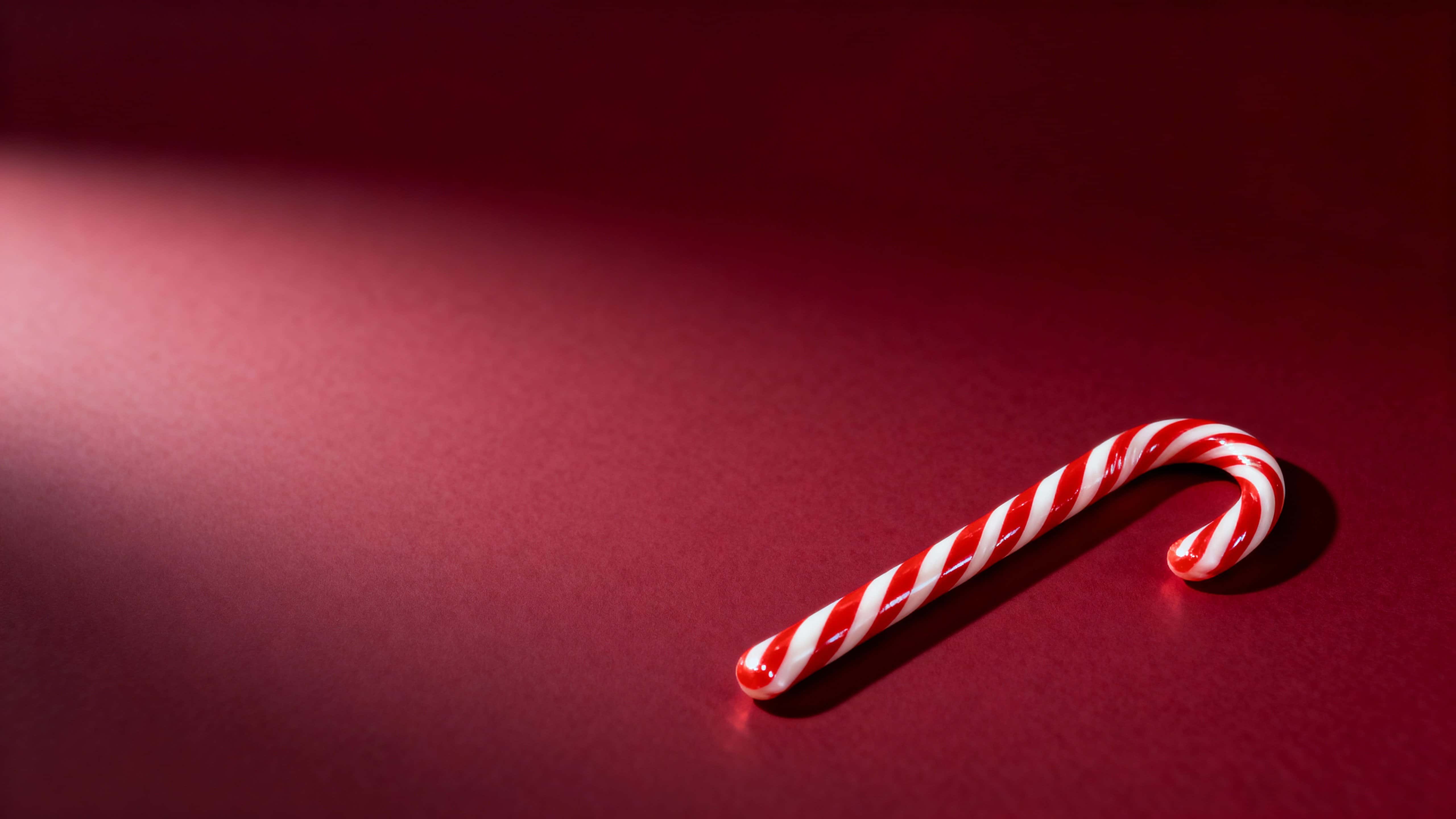 A single glossy red-and-white candy cane lies diagonally in the lower-right of a 16:9 frame against a dark ruby-red gradient background with ample empty upper-left copy space.