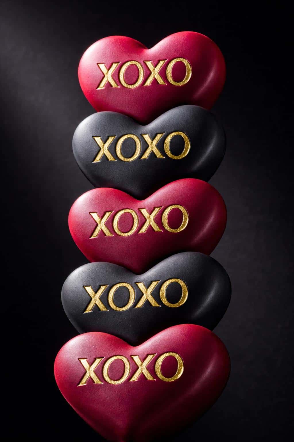 Vertical 2:3 render of stacked ruby and charcoal luxury candy hearts with debossed XOXO and gold-foil inlays, matte-satin finish, and a cold dark background.