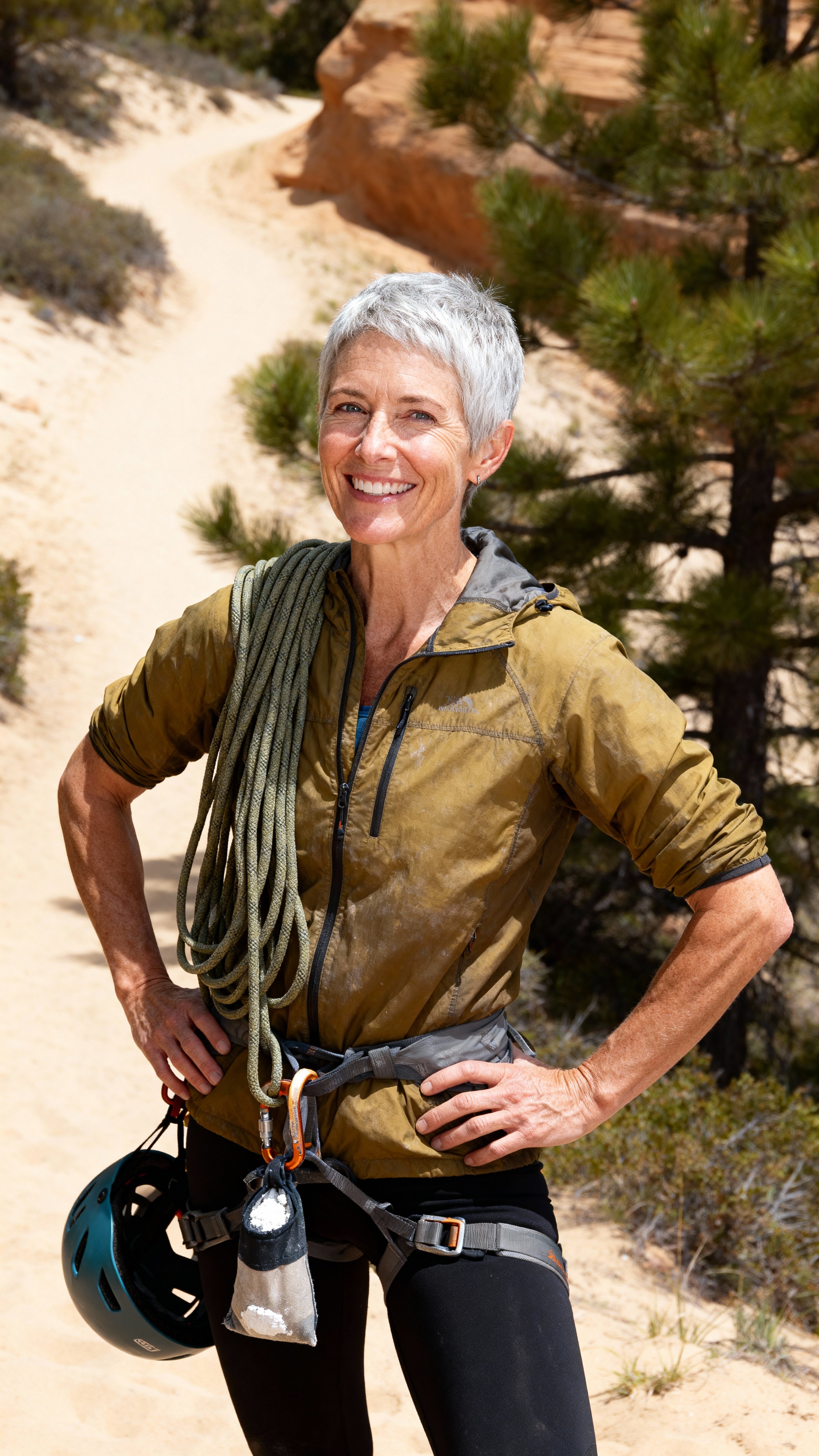 Full-figure portrait of Carolyn, a white climbing coach in her early 70s at a Flagstaff trailhead with coiled rope, chalk bag and helmet, sandy path and soft pines in tones #CBAE88 #2F5A3D #D17C4A #ECEBE7 #2B3B4E.