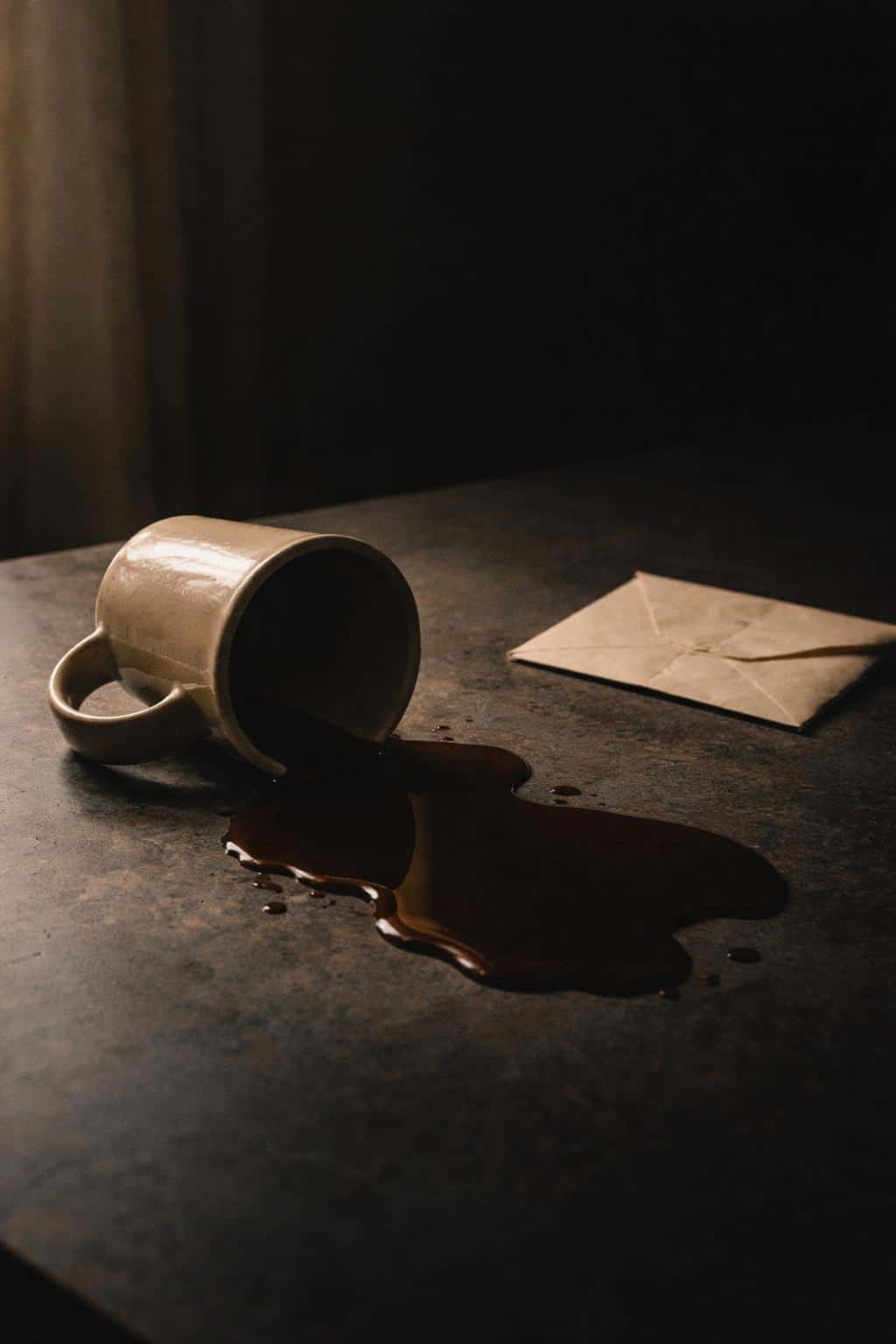 Vertical 2:3 cinematic still-life of a tipped ceramic mug spilling coffee across a dark tabletop beside a folded envelope, minimal and quietly mournful.