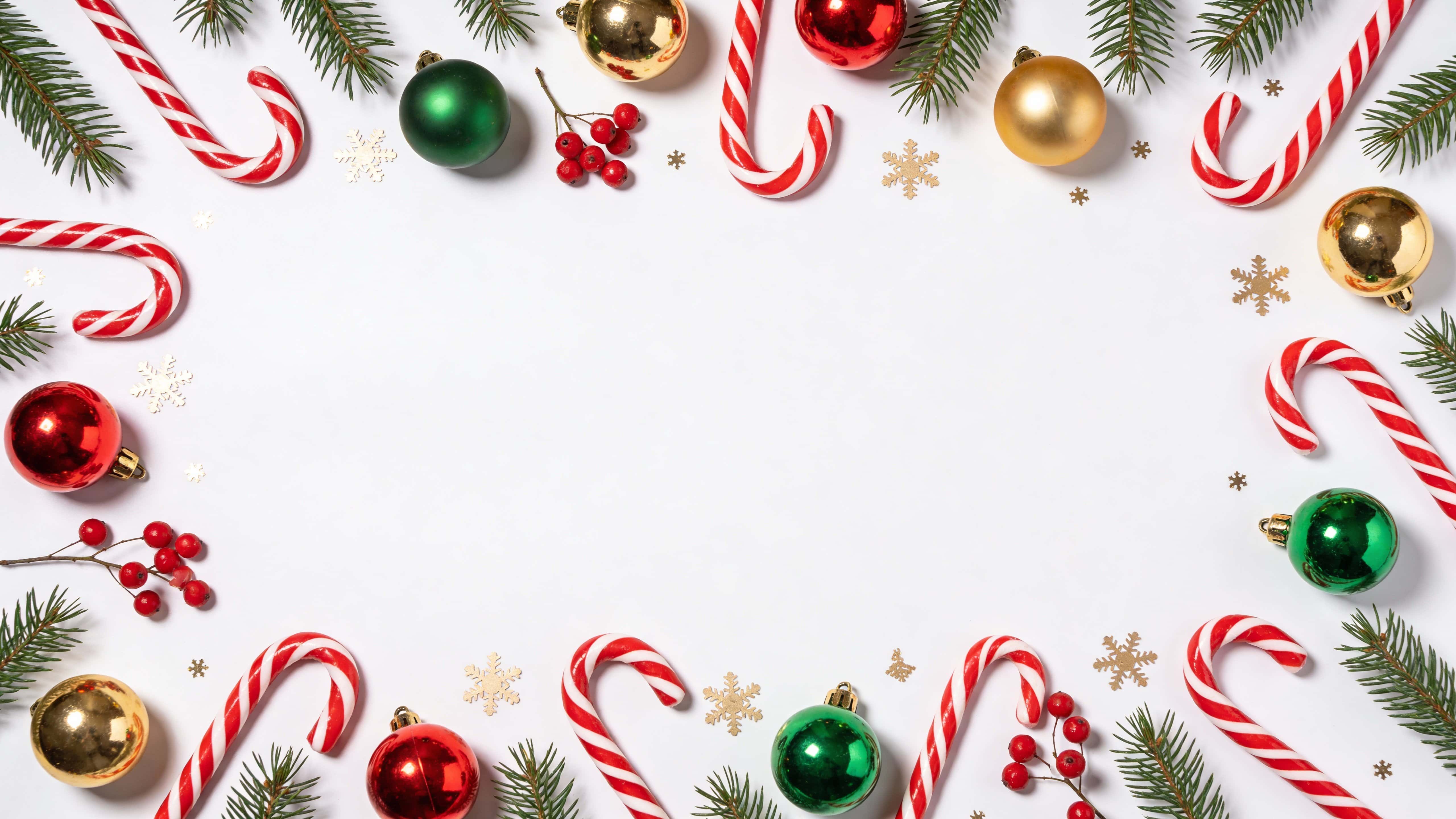 Top-down Christmas flat-lay border on a clean white background with pine branches along the top edge, evenly spaced red-and-white candy canes, red, green and gold baubles, clusters of red berries and light gold snowflake confetti, leaving a