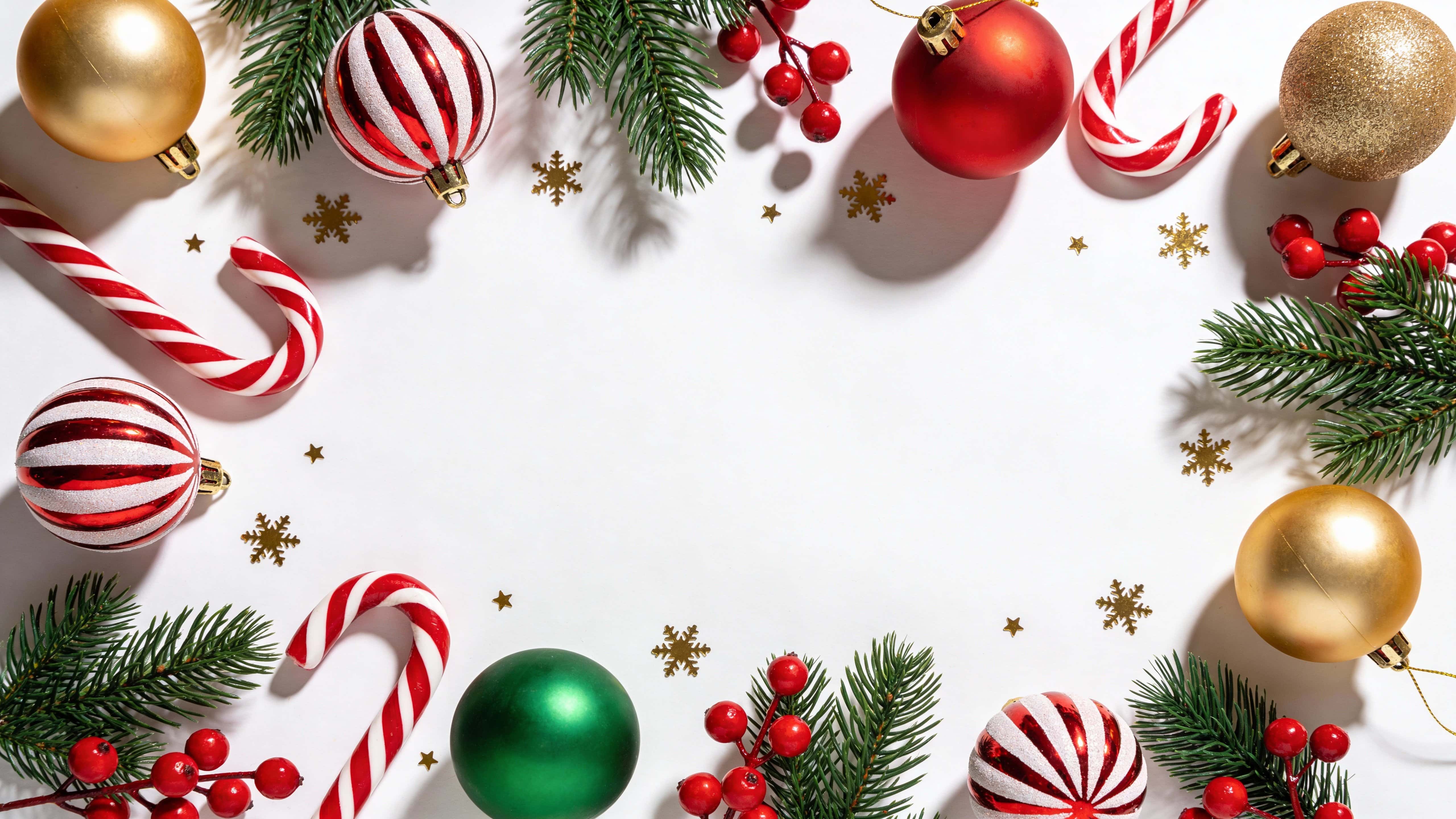 Top-down 16:9 Christmas flat lay border on a white background showing fresh pine branches, diagonal red-and-white candy canes, red, green and gold baubles, red berries and gold snowflake confetti.