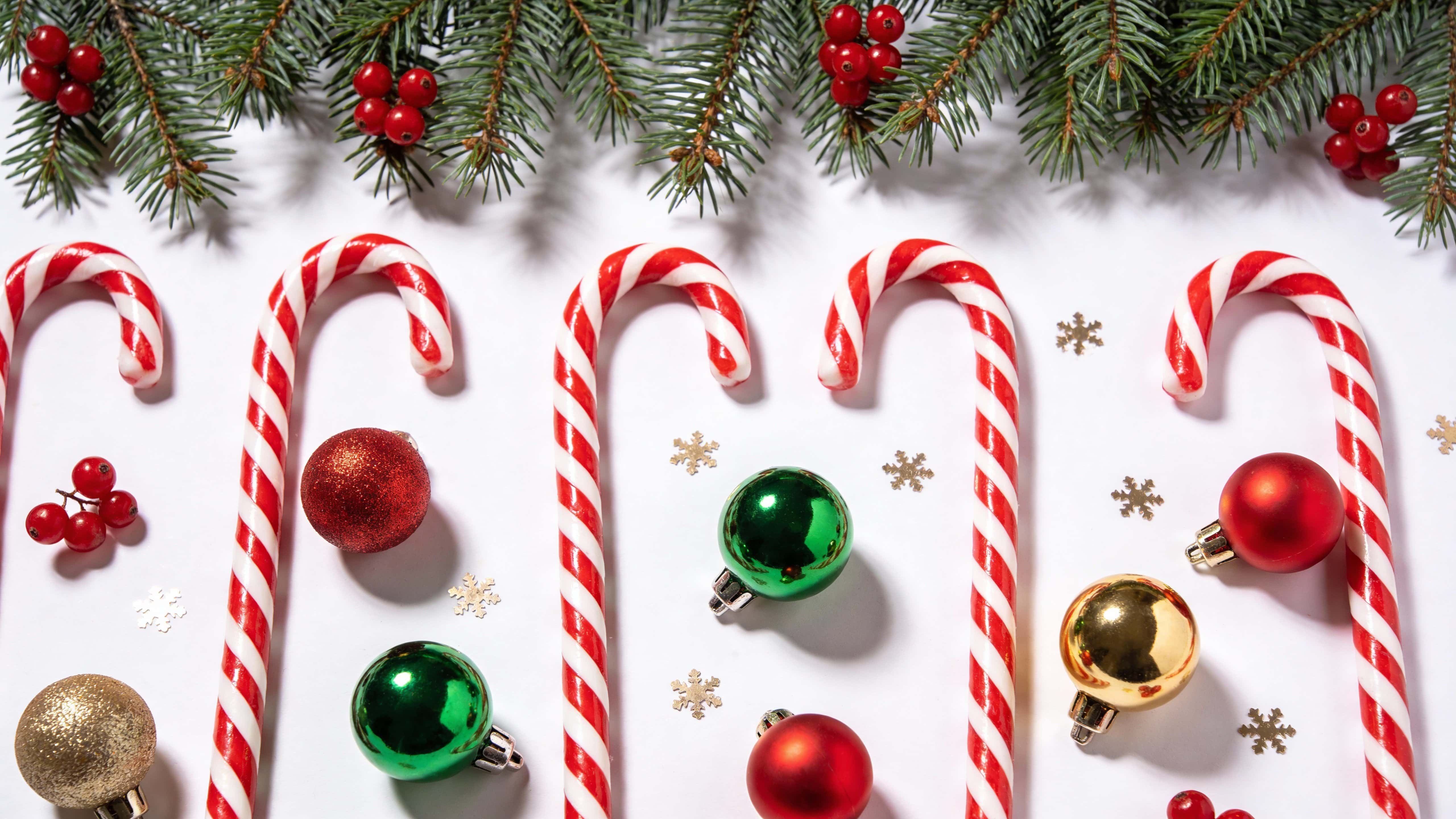 Top-down Christmas flat lay border on a white background with pine branches along the top, evenly spaced red and white candy canes, glossy red, green and gold baubles, red berries and gold snowflake confetti.