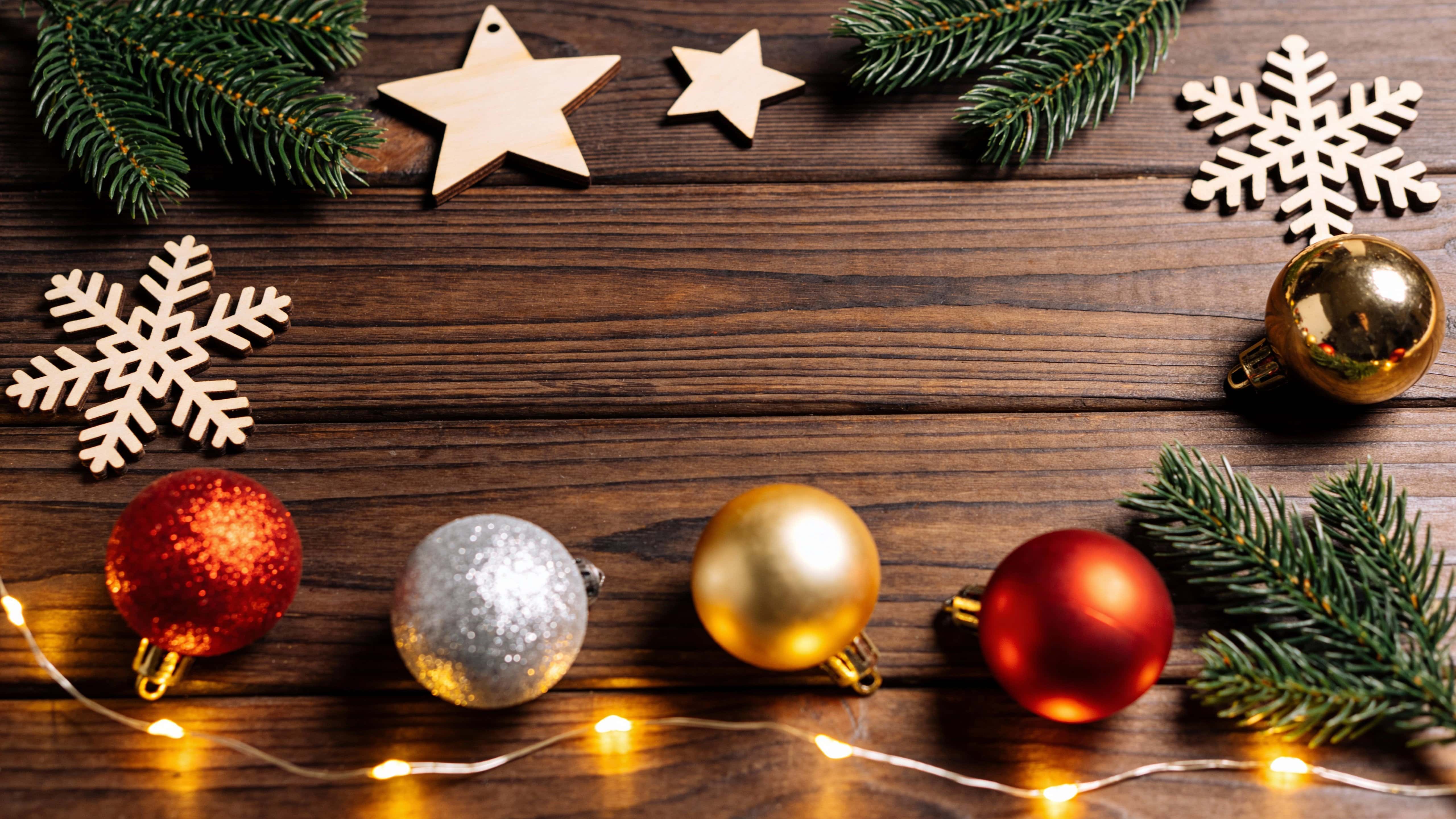 Top-down 16:9 rustic Christmas flat lay on dark brown horizontal wooden planks with pine branches, wooden stars and snowflakes, red/gold/silver baubles and warm fairy lights framing top and bottom, center left empty for copy space in a cozy