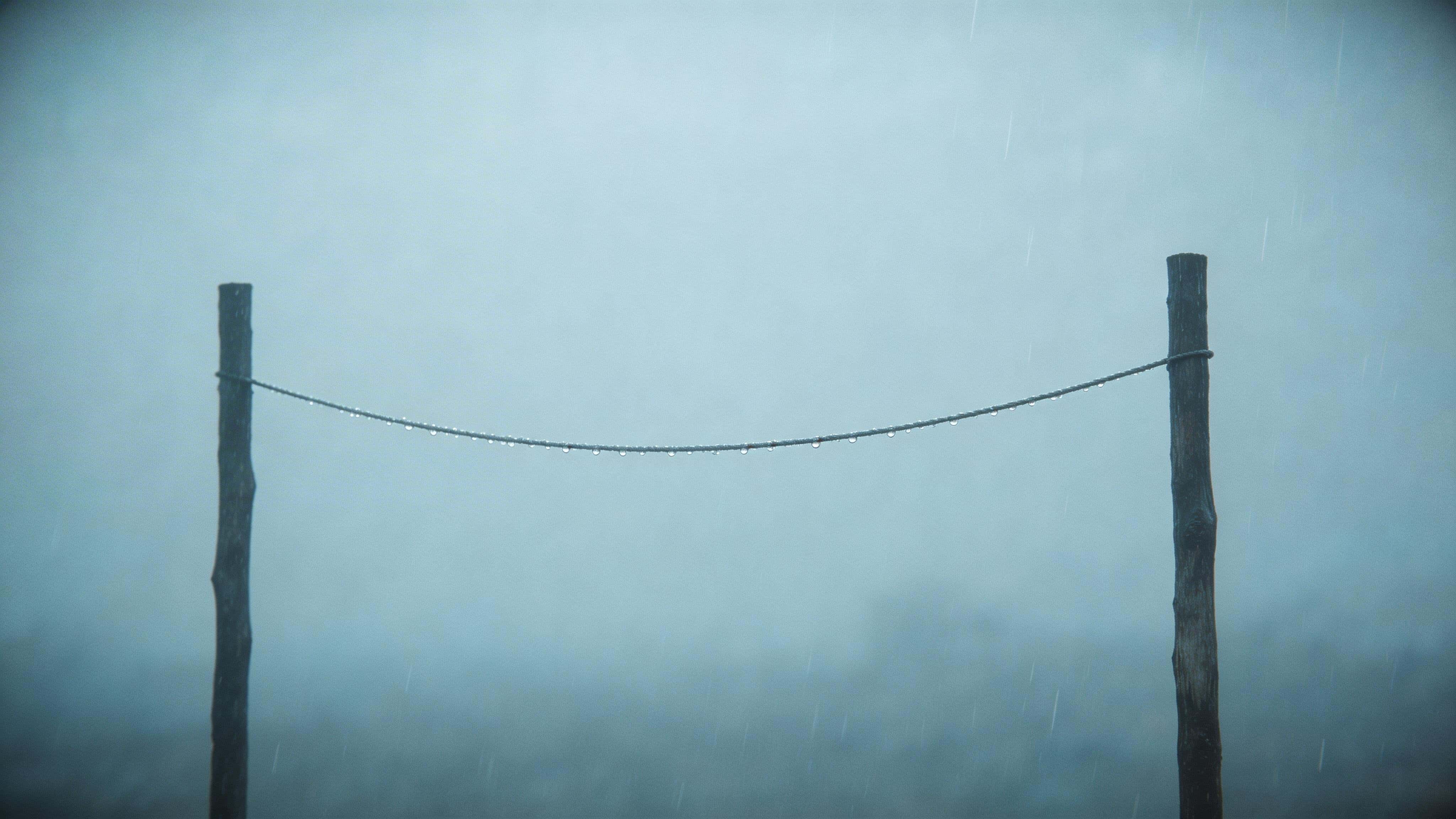 Minimal cinematic 16:9 wallpaper of an empty clothesline between two posts in steady rain, muted greys and cool blues with soft mist, quiet and unoccupied mood.