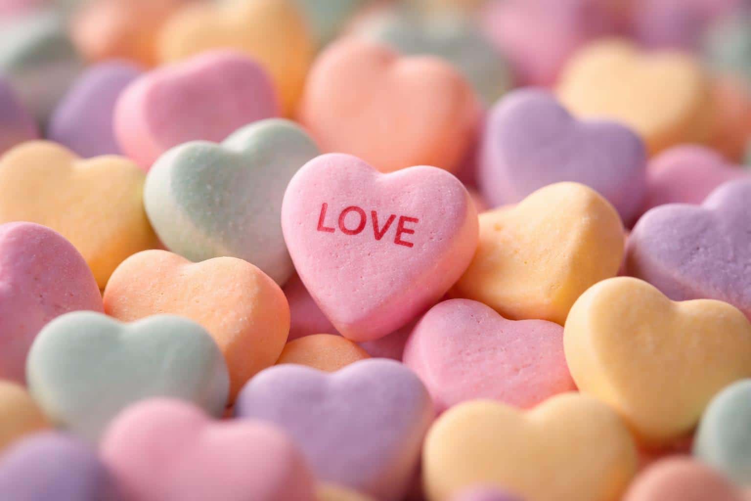A cinematic macro of a wide 3:2 pile of pastel conversation hearts with a central 'LOVE' heart in red, matte chalky texture, soft natural light, and shallow depth of field.