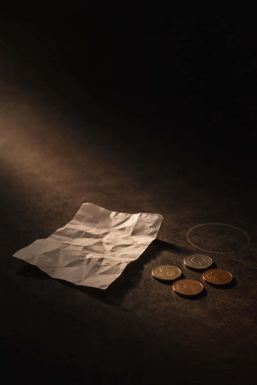 Vertical 2:3 cinematic tabletop still-life of a crumpled paper receipt and loose coins in the lower third, warm side window light, deep shadows, and subtle film grain.