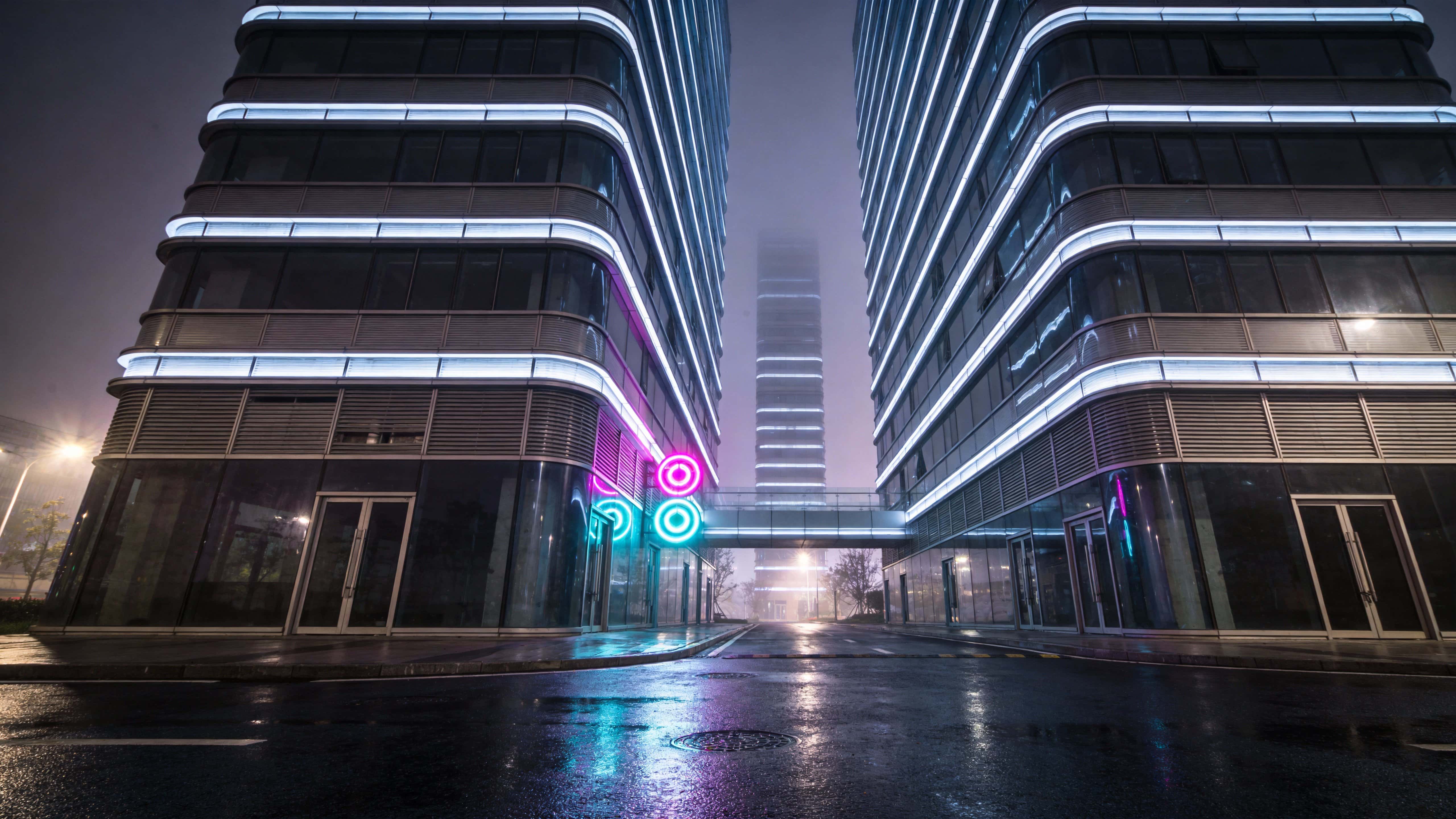 Low-angle, polished architectural night photograph of a cyberpunk city in 16:9, with a dense wet avenue and vertical towers arranged for dramatic wallpaper use.