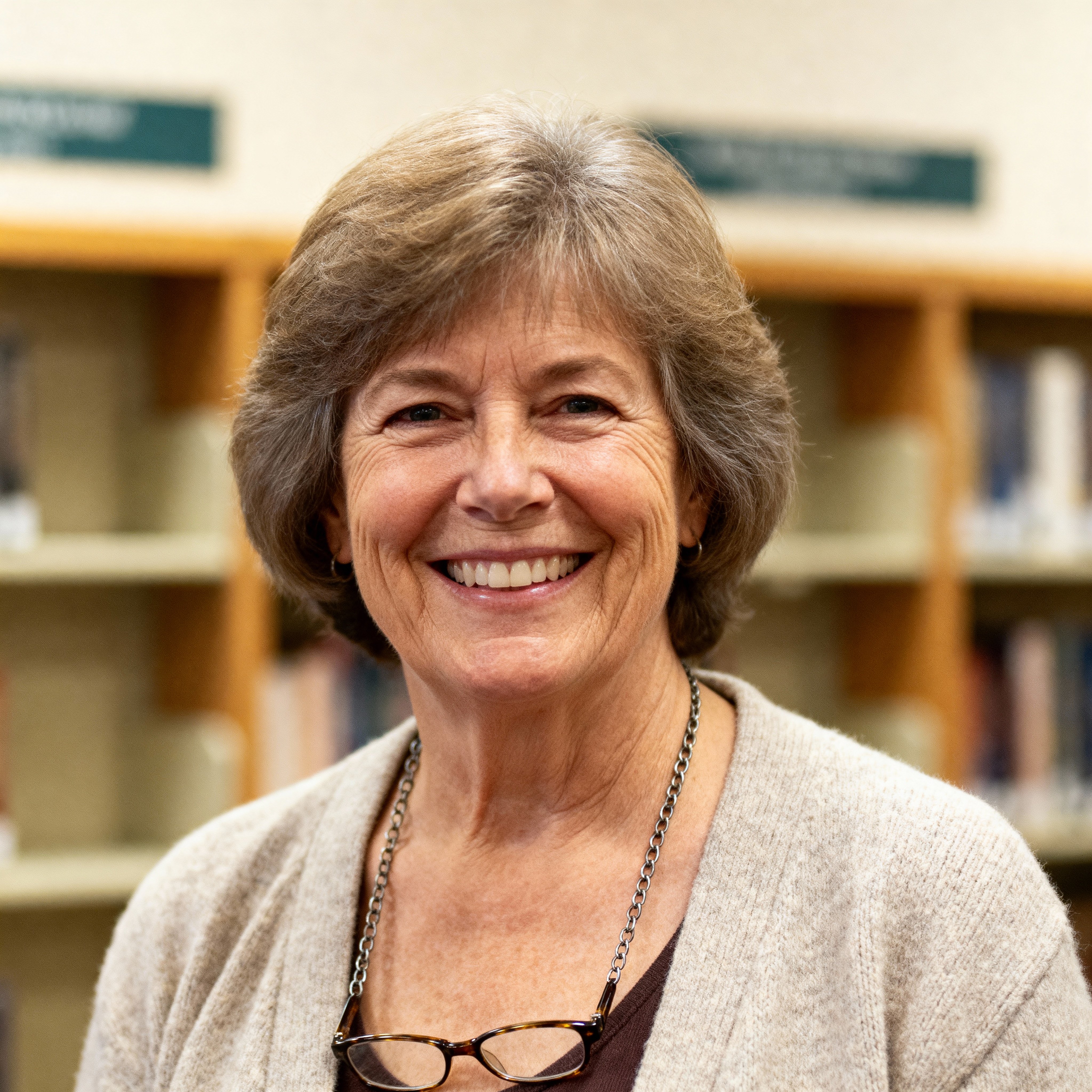 Headshot of Elaine Hart, a 63-year-old white Appalachian librarian in a cream cardigan with a sage scarf and soft-gold accents, glasses on a chain, warm smile, gentle flattering light, neutral blurred stacks with muted-blue and warm-brown t