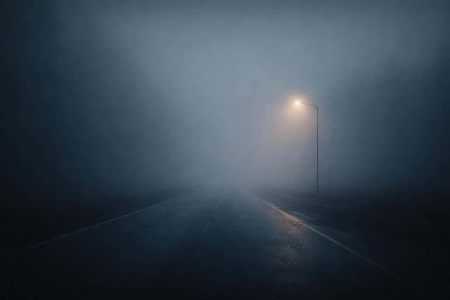 Minimal cinematic 3:2 landscape of an empty road vanishing into dense morning fog, with a lone off-center streetlight casting a soft warm halo on damp pavement.