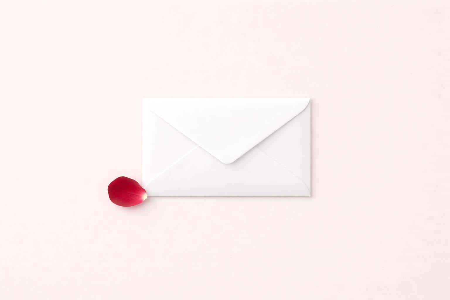 Minimalist 3:2 landscape flat lay of a pristine white envelope on a soft blush-pink background with a single red rose petal and ample negative space.