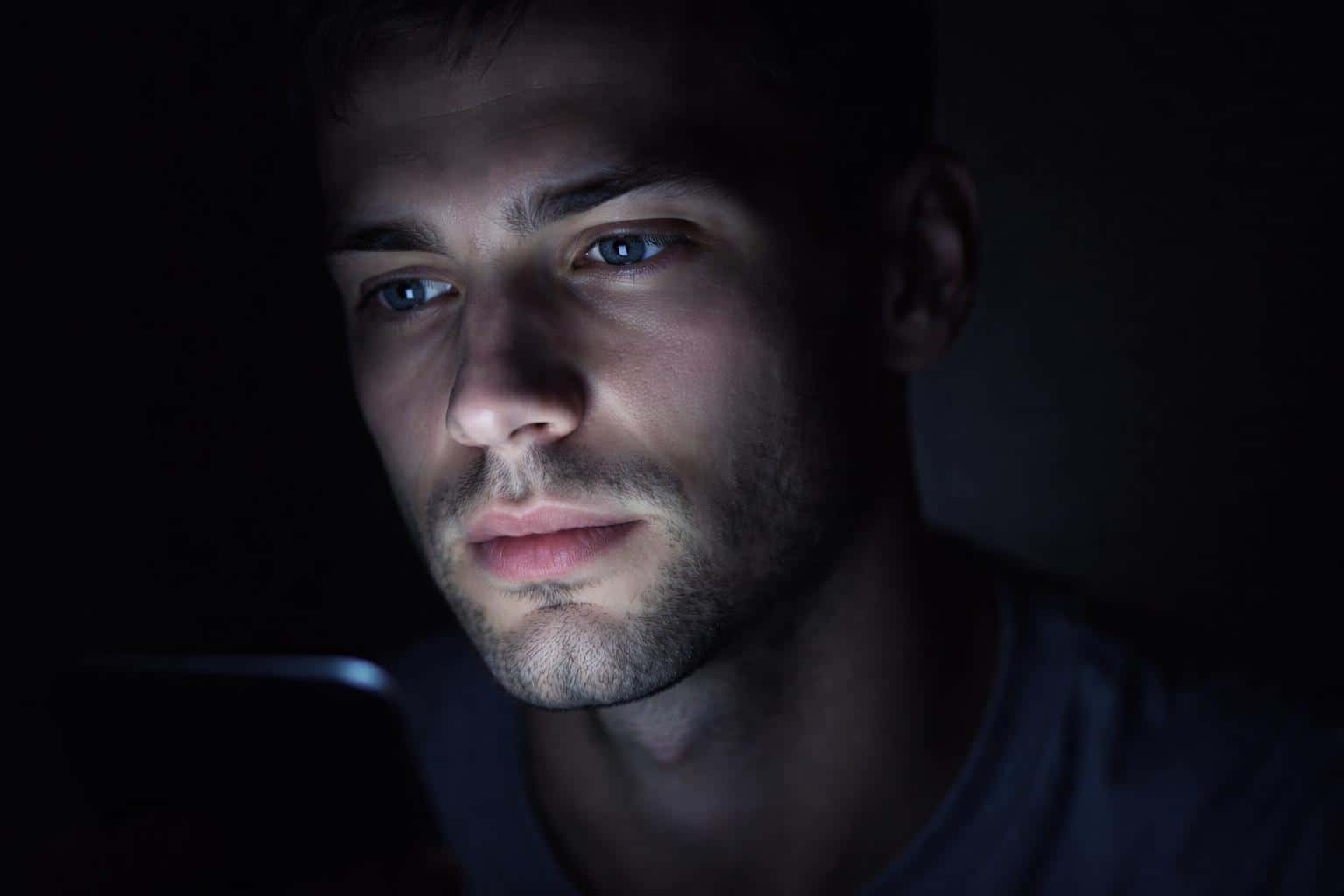 Close-up portrait of a face lit only by a phone screen in dark surroundings, a moody photographic study intended as a poster-cover with cinematic contrast.