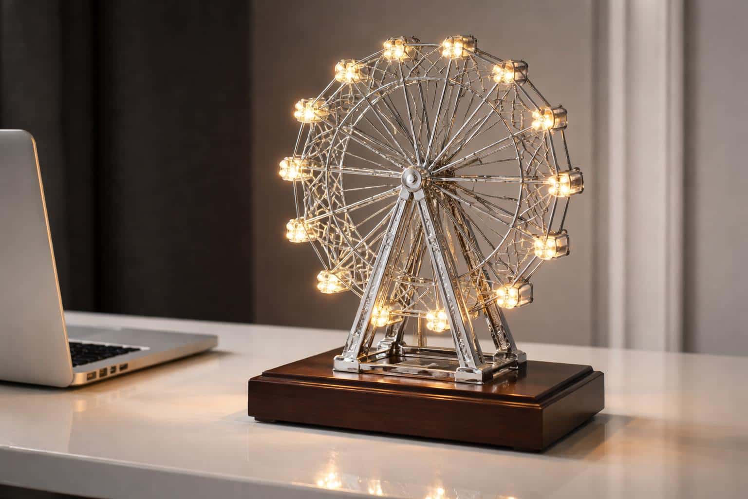 A realistic product photograph of a silver metal ferris wheel lamp with glowing warm LED lights, positioned on a wooden base atop a white desk surface.