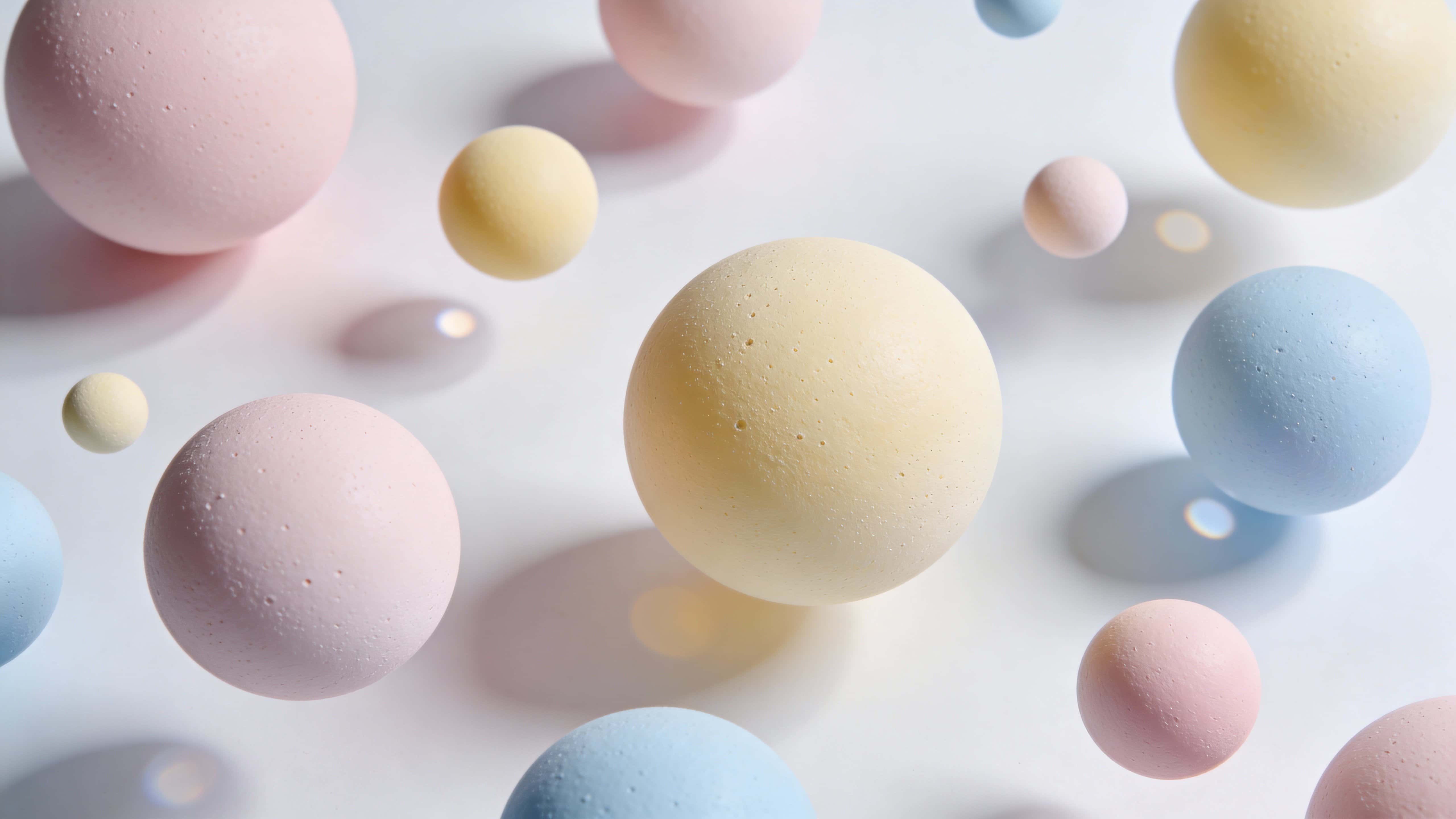 Overhead 16:9 view of floating spheres scattered across a shallow plane with varied sizes and edge crops, matte micro-texture, soft shadows, muted pinks, pale yellow, soft blue, and cool-white fill.