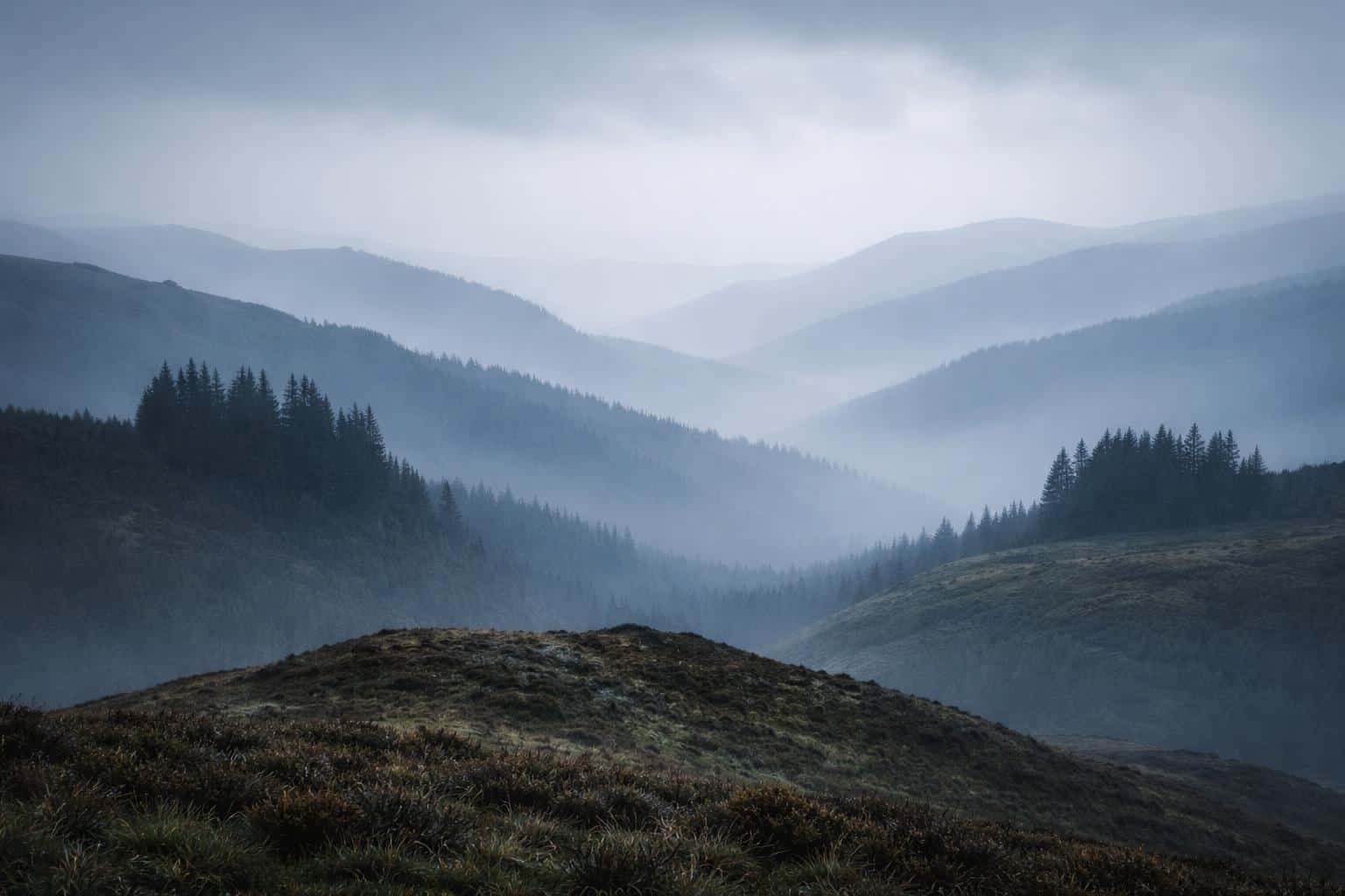 Fog-covered hills layered into the distance in a 3:2 wide landscape photograph, with soft diffuse light, cool muted tones and deep tonal separation for wallpaper.