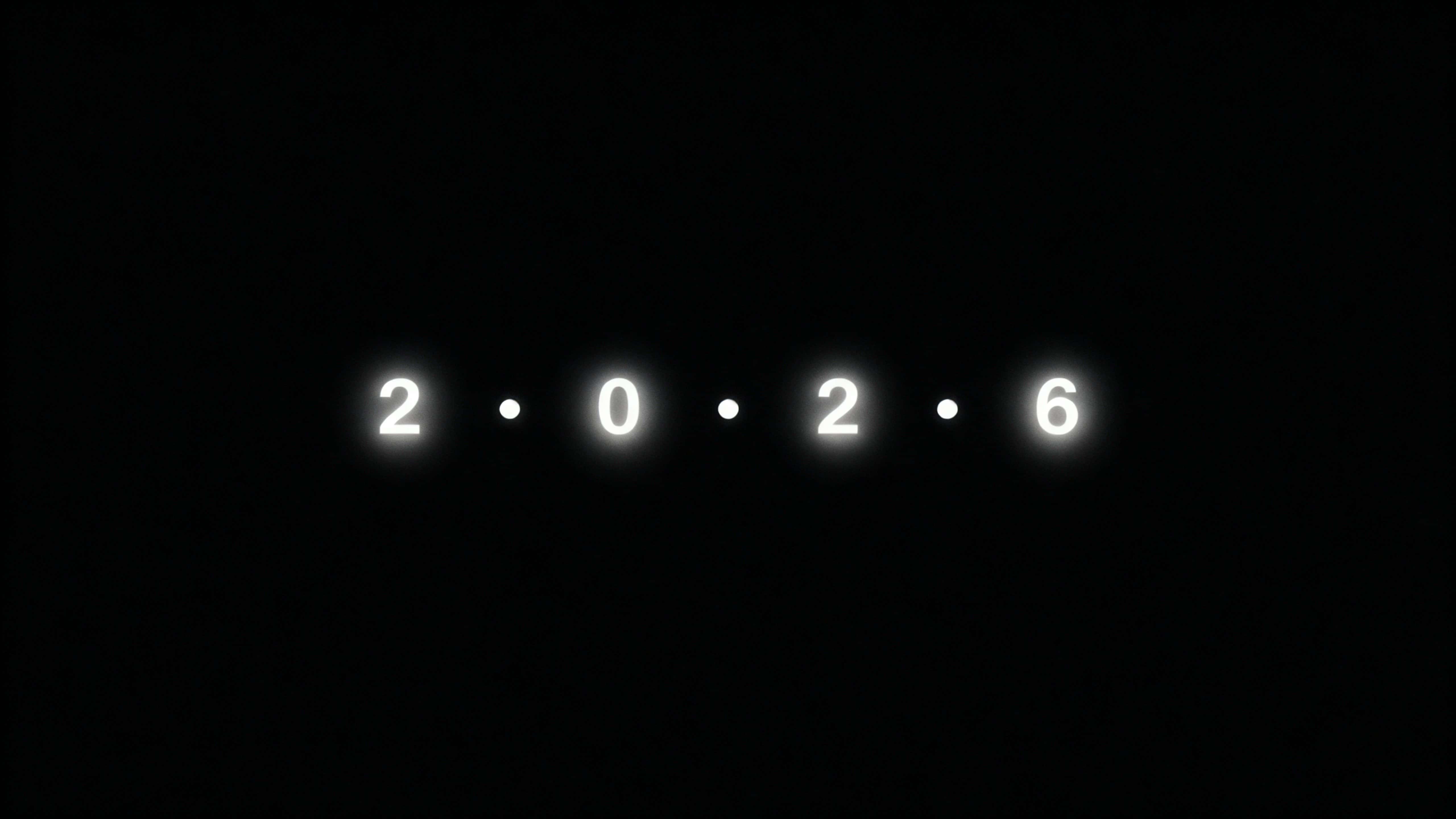 Four small luminous dots float in a perfect horizontal line within a 16:9 frame, each pulse briefly forming a tiny numeral — 2 • 0 • 2 • 6 — against deep black negative space.