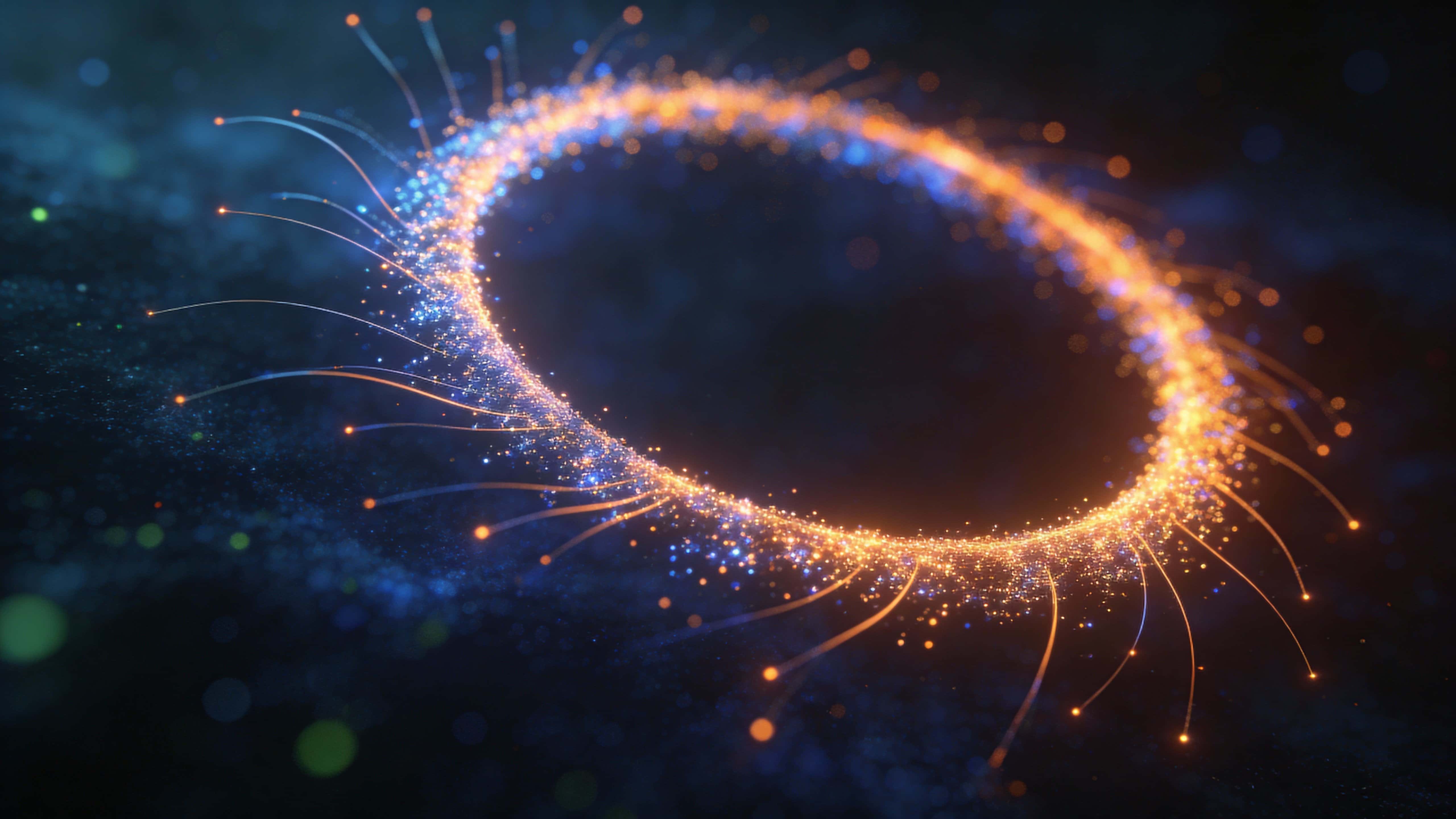 A nearly perfect, tilted ring of thousands of blue, orange, and soft-green particles shimmers in deep darkness with short filaments radiating outward and a calm inner void.