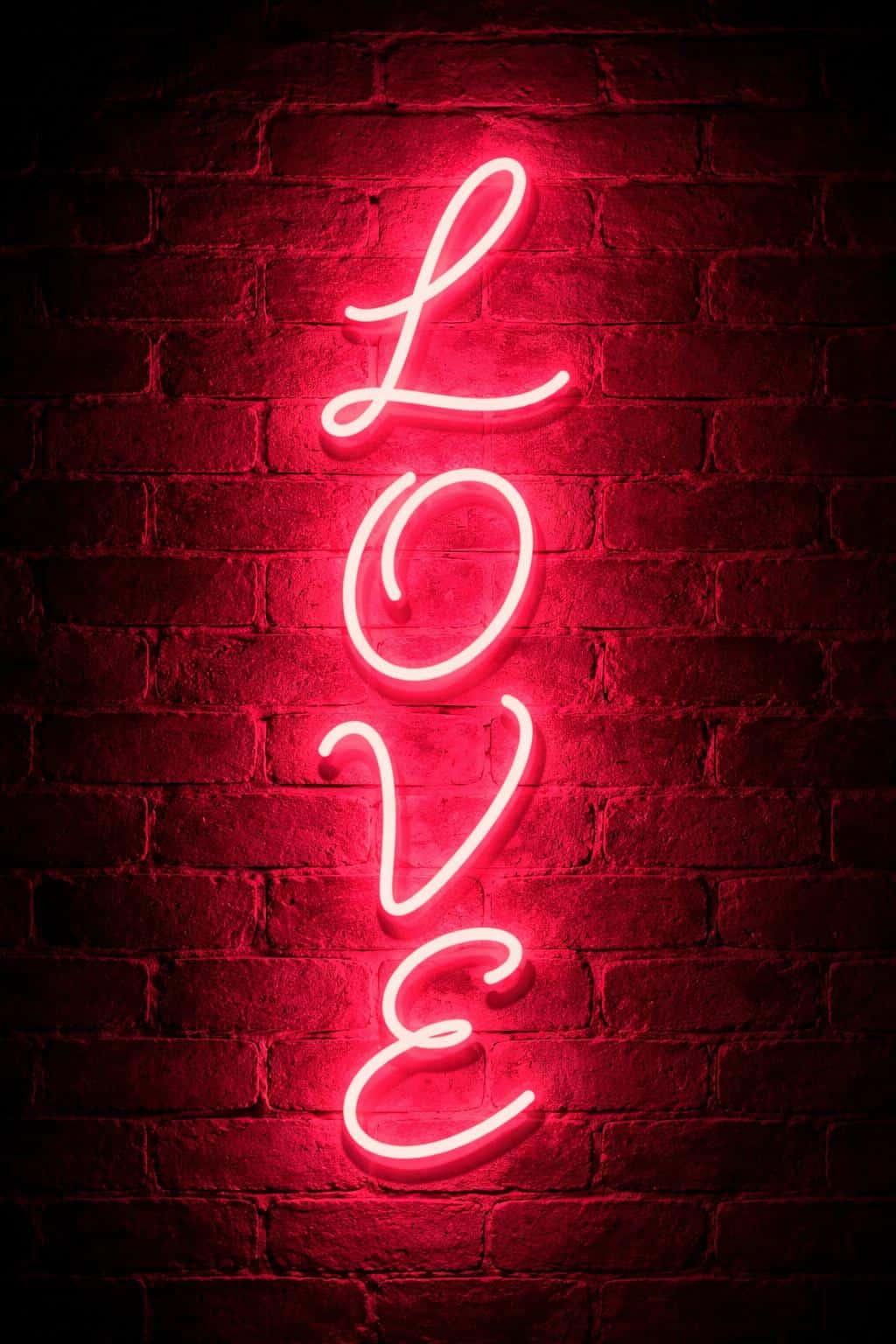 Cinematic vertical 2:3 night scene of stacked cursive 'L O V E' neon on a dark red brick wall, the intense pink halo saturates mortar and rough brick texture for an intimate, urban mood.