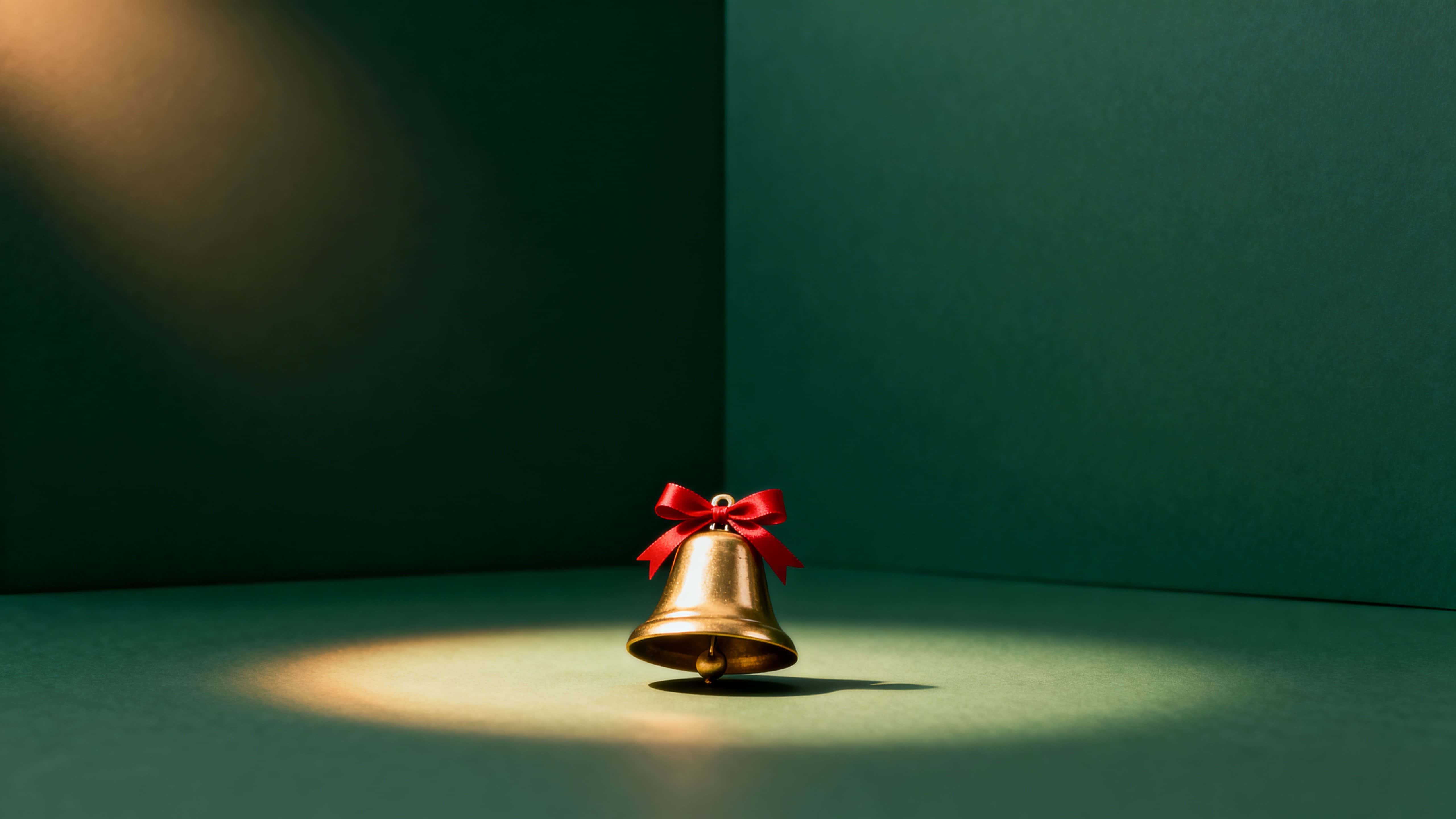 A wide 16:9 Christmas card showing a single smooth golden bell with a red ribbon centered near the bottom, set against a deep evergreen gradient halo and open upper copy space.