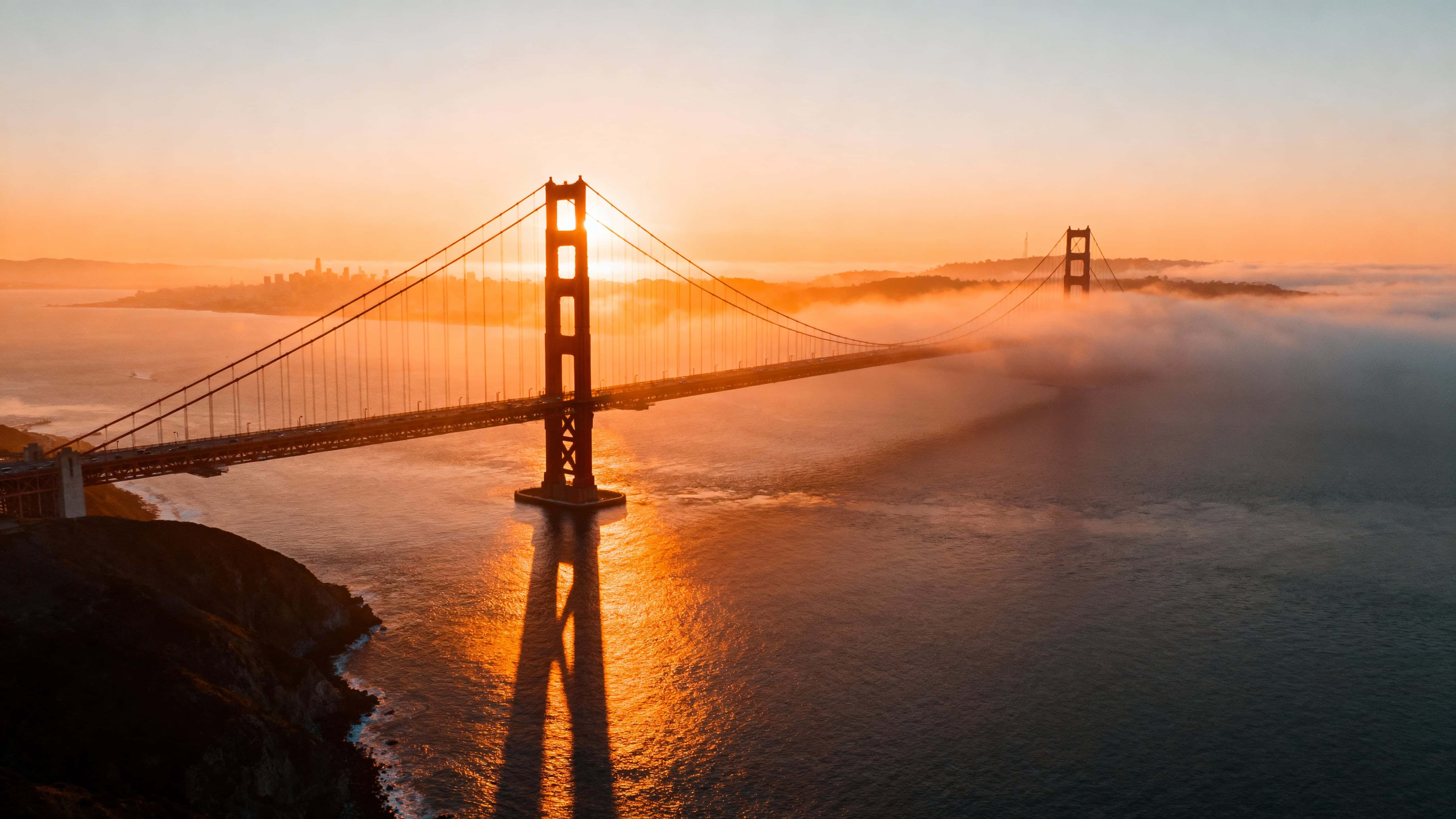 Aerial 16:9 composition of the Golden Gate Bridge at sunrise, orange sunlight piercing morning mist with long shadows and calm bay reflections in ultra-realistic warm tones.