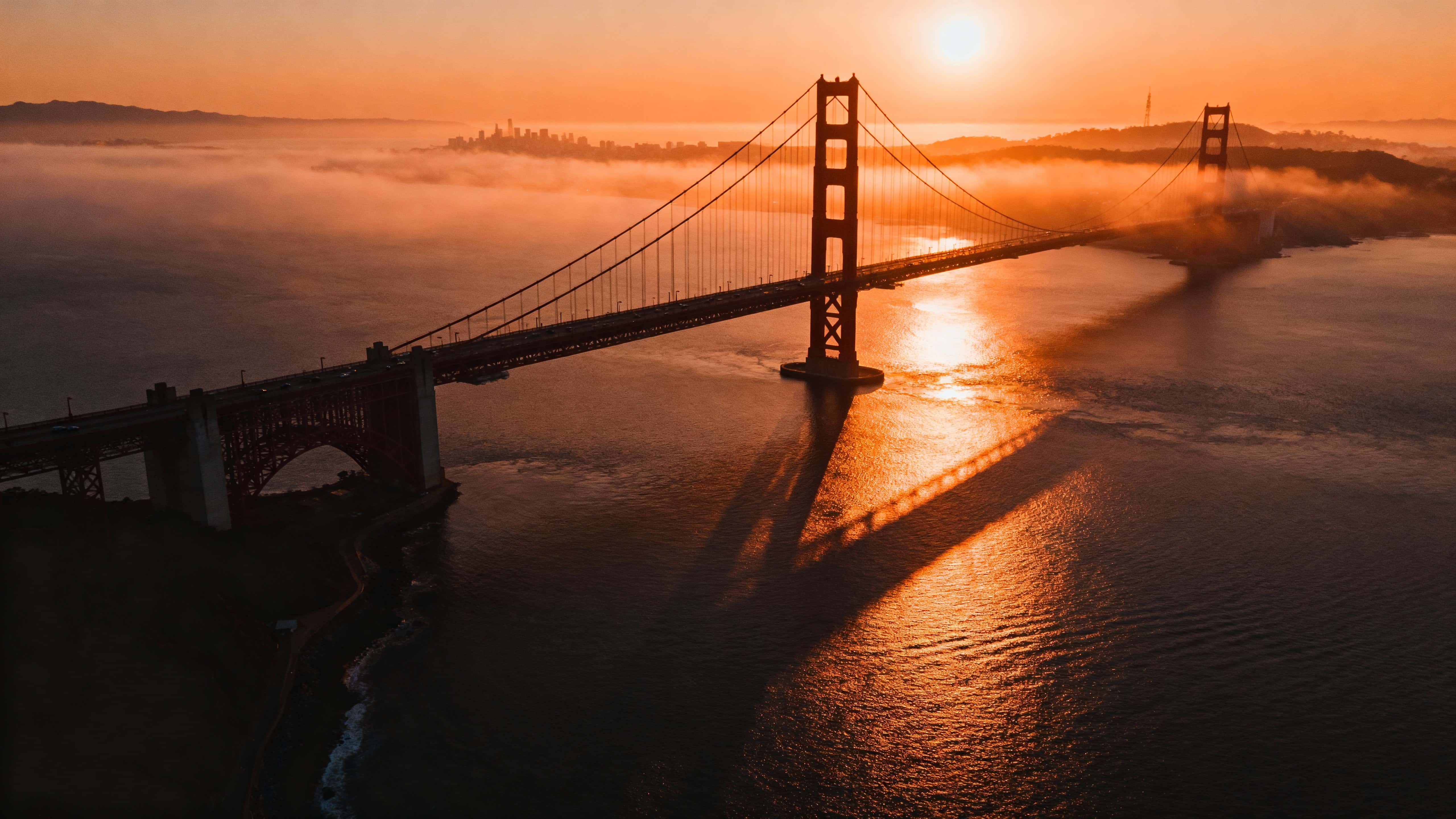 Aerial 16:9 composition of the Golden Gate Bridge at sunrise, orange sunlight piercing morning mist with long shadows and calm bay reflections in ultra-realistic warm tones.