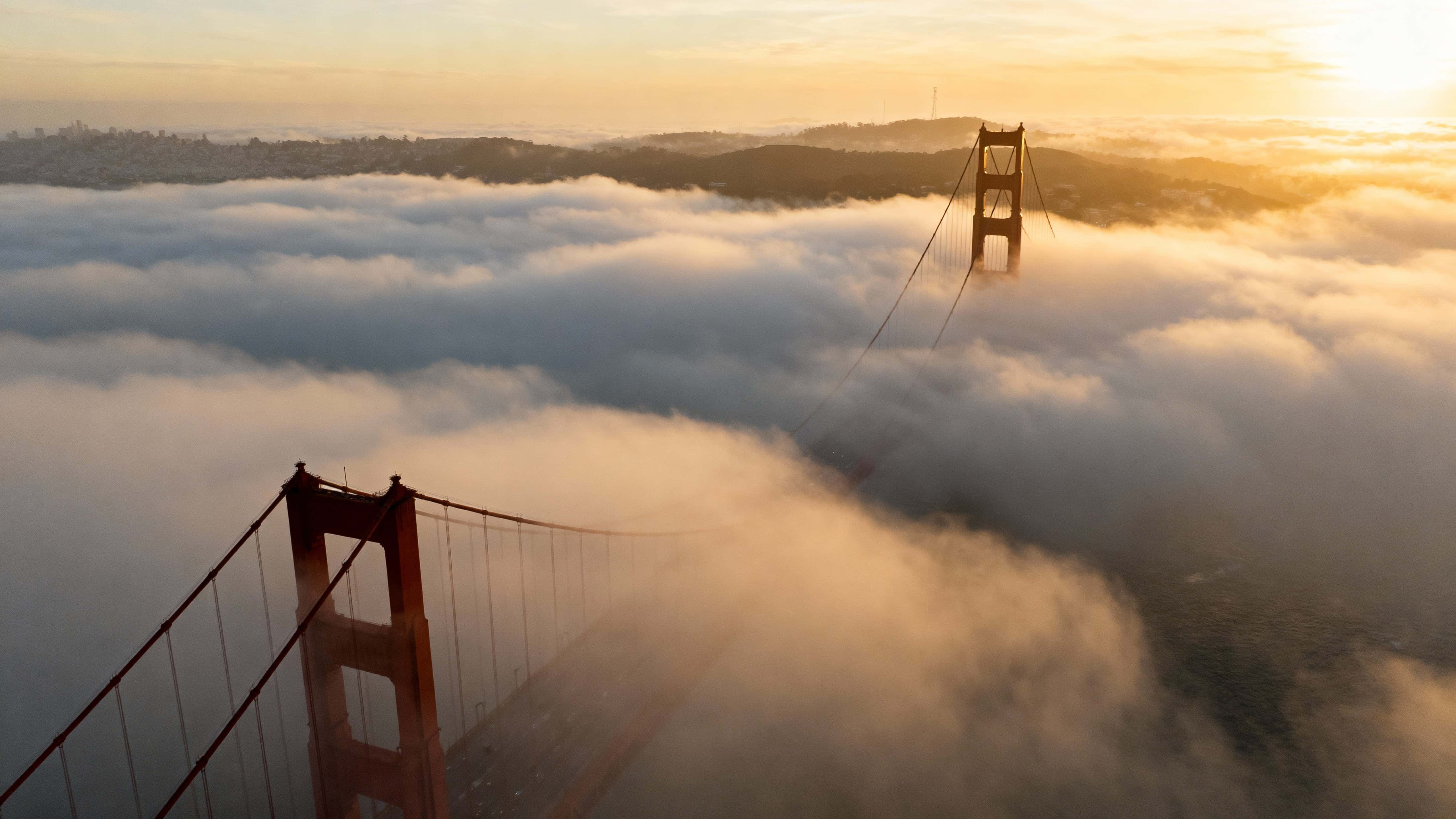 Drone-eye view of Golden Gate Bridge towers piercing a thick rolling fog at dawn, tops visible above clouds with warm golden hues and soft scattered light in a cinematic 16:9 frame.