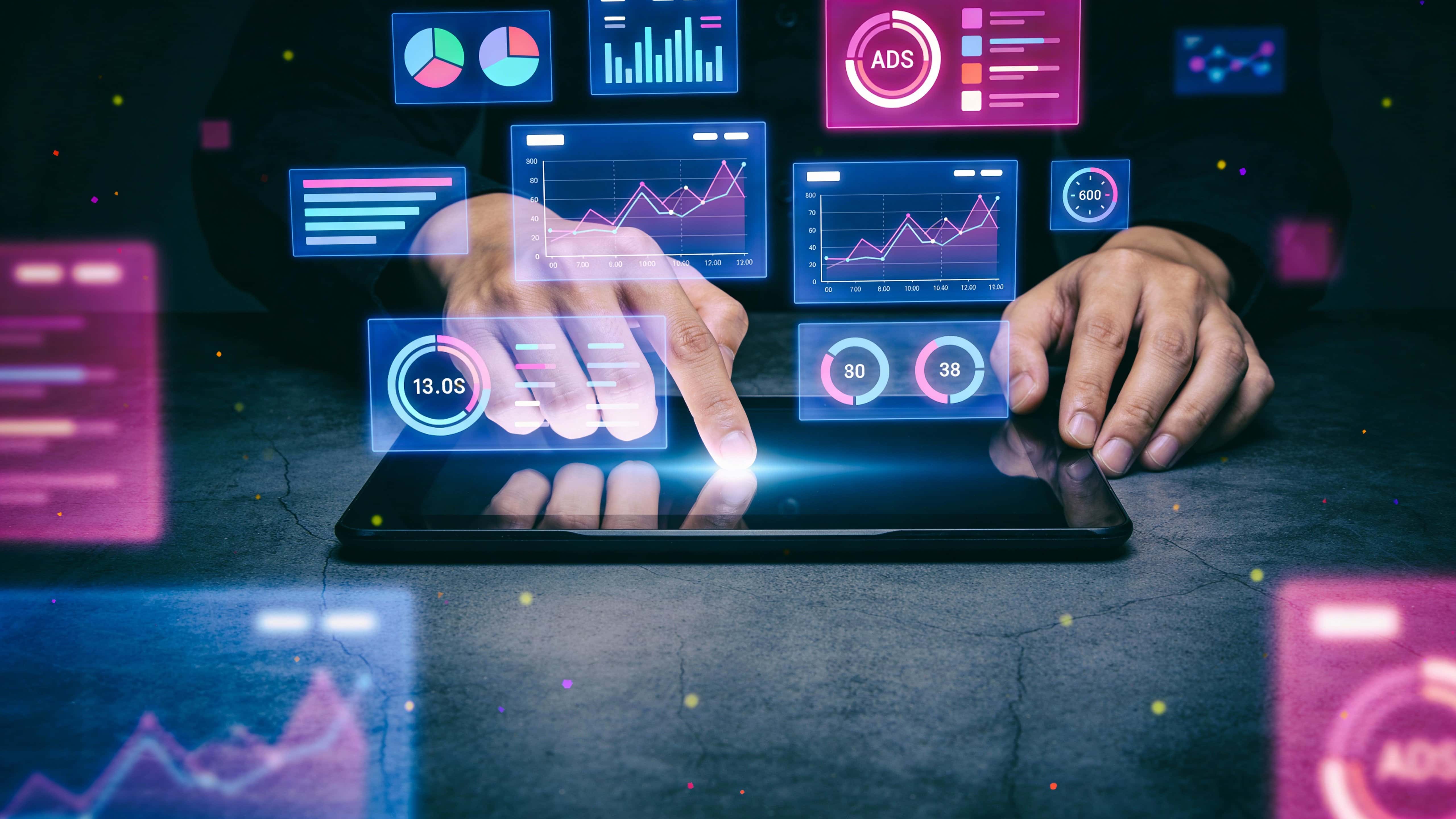 Dark futuristic workspace with hands interacting with a digital tablet beneath floating neon-blue and magenta holographic dashboards, charts and glowing UI tiles softly illuminating the hands and table.