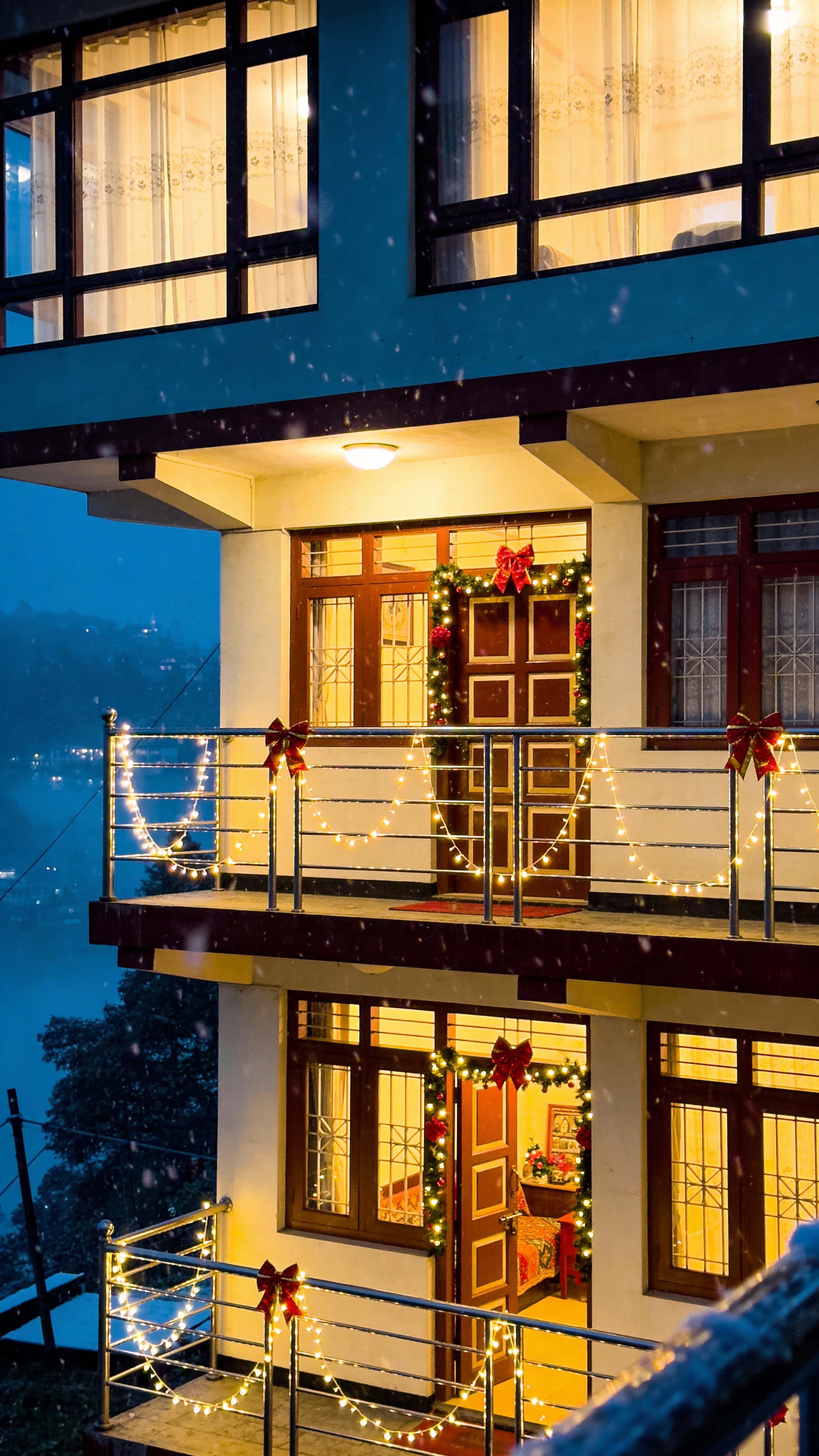 A cozy hillside homestay in Darjeeling on a Christmas night with warm interior glow, soft fairy lights around windows, gentle mist and falling snow-like particles, and red-gold festive accents.