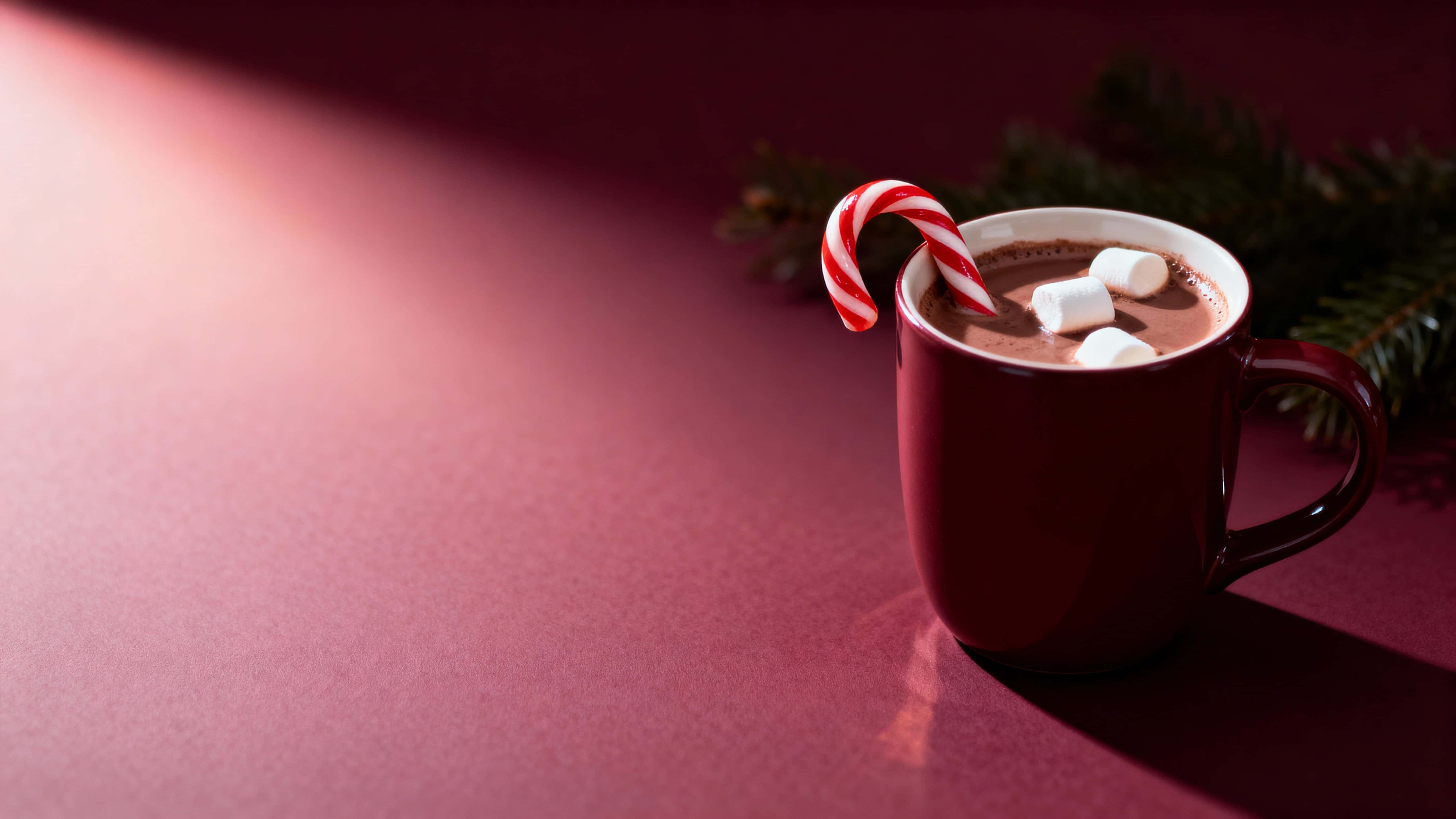 A 16:9 Christmas background showing a cozy ceramic mug of hot chocolate with a hooked candy cane and floating marshmallows on the right third, set against a soft burgundy gradient with faint pine branches and generous left-side copy space.