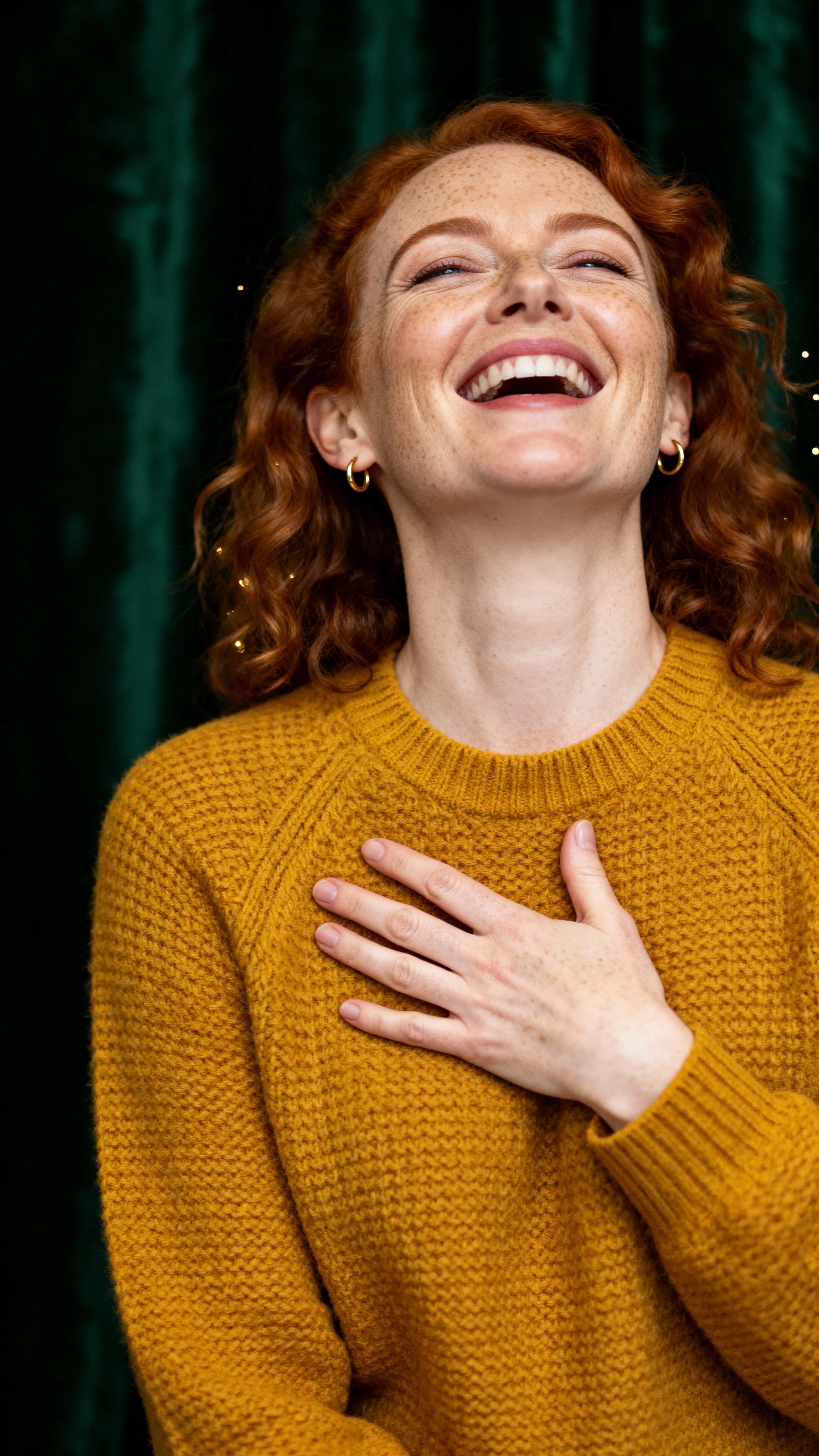 Warm 9:16 studio portrait of a 35-year-old Irish woman with alabaster freckles and loose copper curls, mid-torso framed and laughing in a mustard sweater against a dark forest-green backdrop.