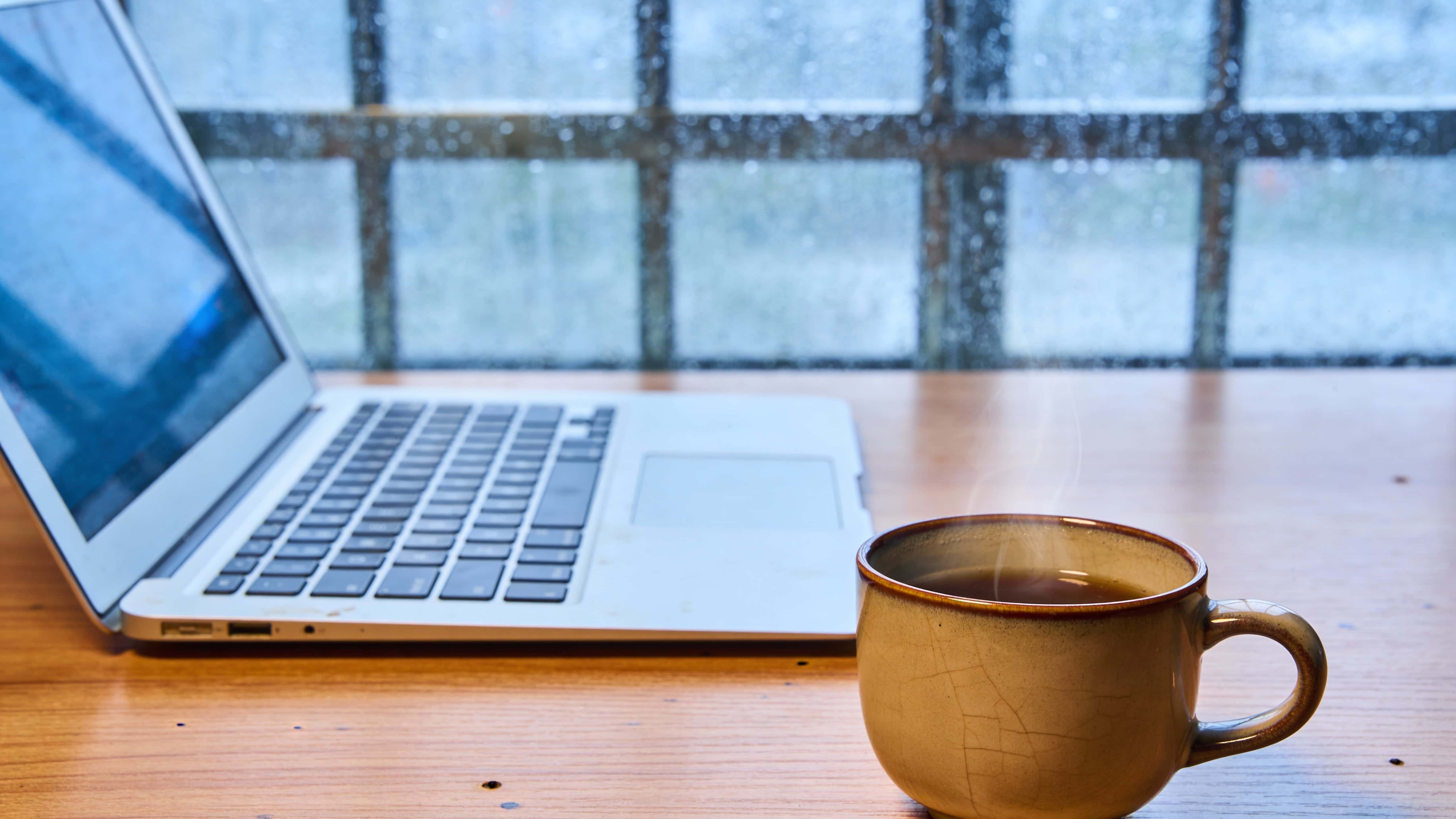 Cozy 16:9 lifestyle photo of a laptop and steaming coffee on a wooden desk beside a rain-speckled window, rendered with soft diffuse light and moody atmosphere.