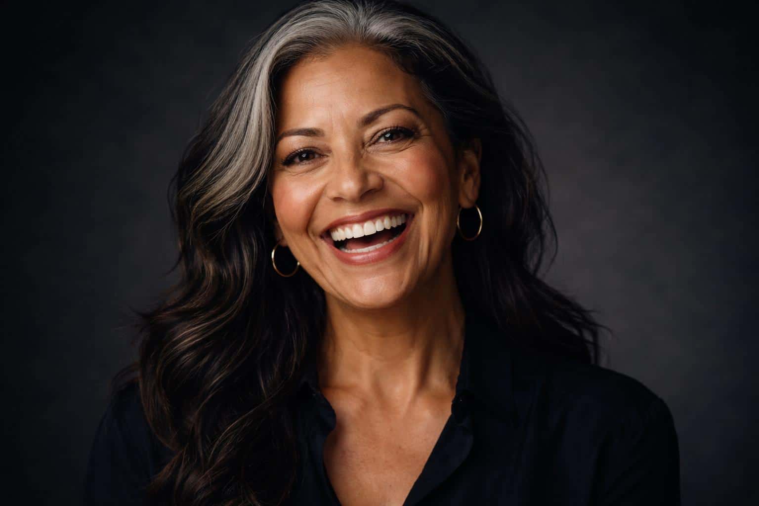 Cinematic 3:2 editorial studio headshot of a joyful early-60s Latina woman with silver-streaked dark hair and warm tan skin, laughing into a genuine smile against a deep charcoal backdrop.