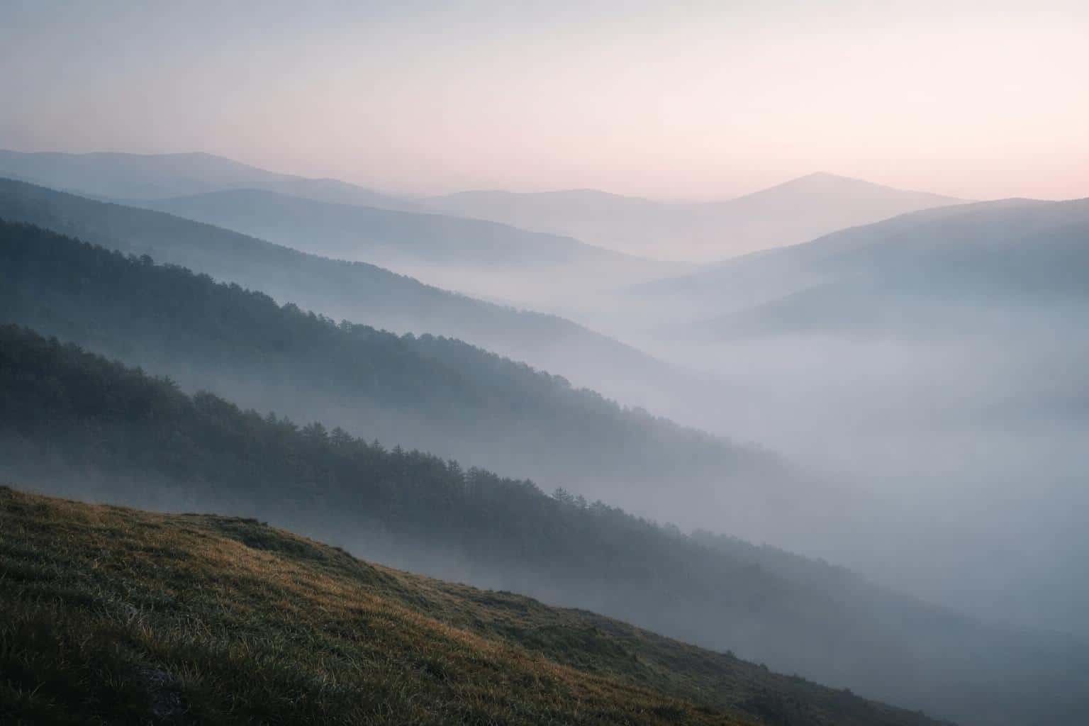 Layered hills shrouded in soft morning mist composed as a 3:2 landscape photograph with tonal depth, delicate pastel shifts and expansive negative space suitable for wallpaper.