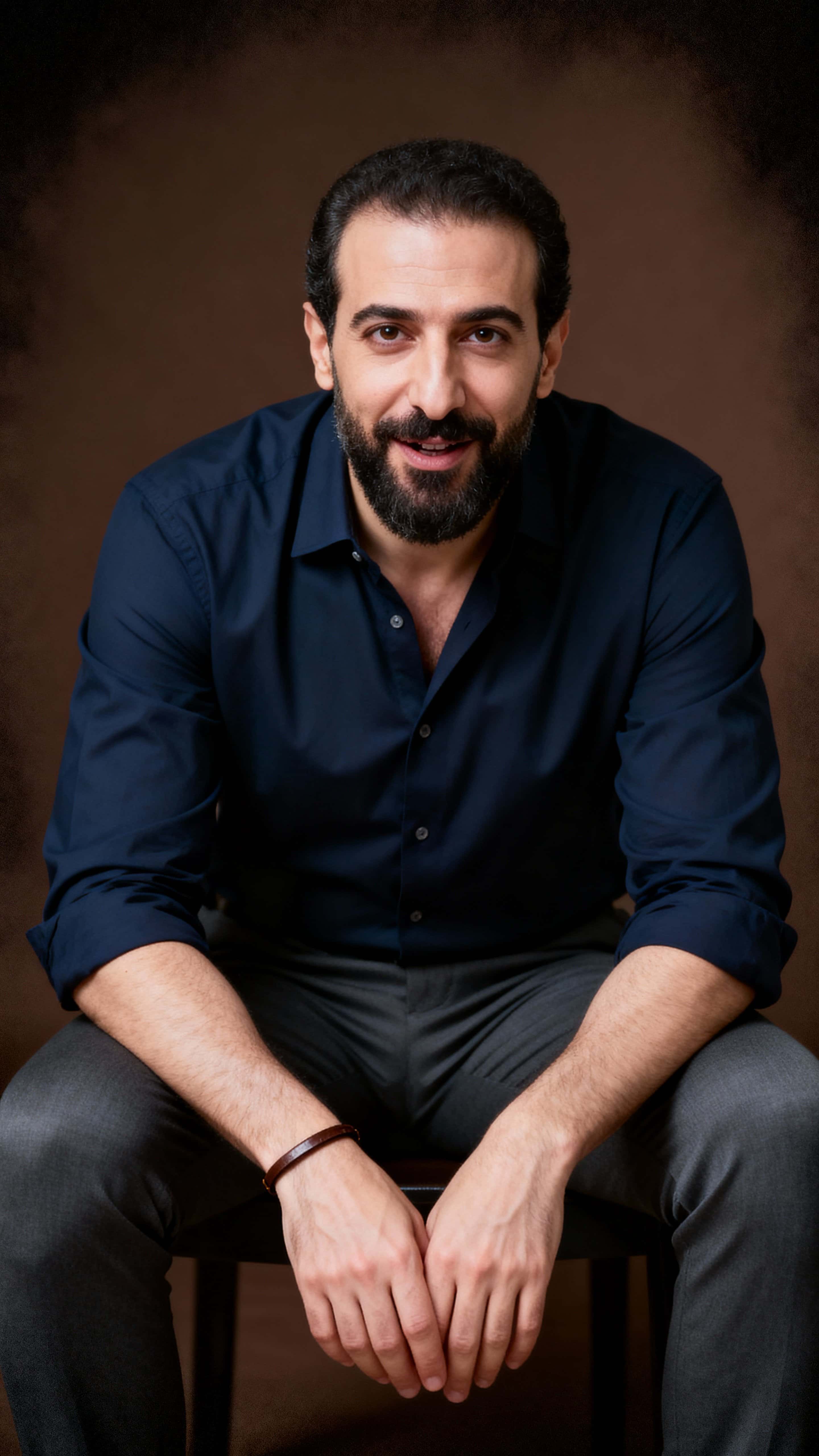 A thoughtful studio portrait of a 40-year-old Lebanese man seated off-center in a 9:16 frame, leaning forward with clasped hands and a conspiratorial gaze against a smoky espresso-brown background.