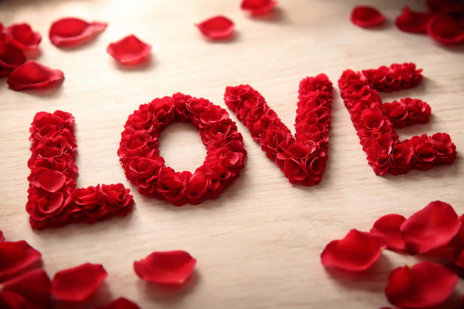 Cinematic macro of the word LOVE formed from hundreds of fresh red rose petals on a light wood surface in a centered 3:2 landscape with soft side-window light and scattered petals for a classic, romantic mood.