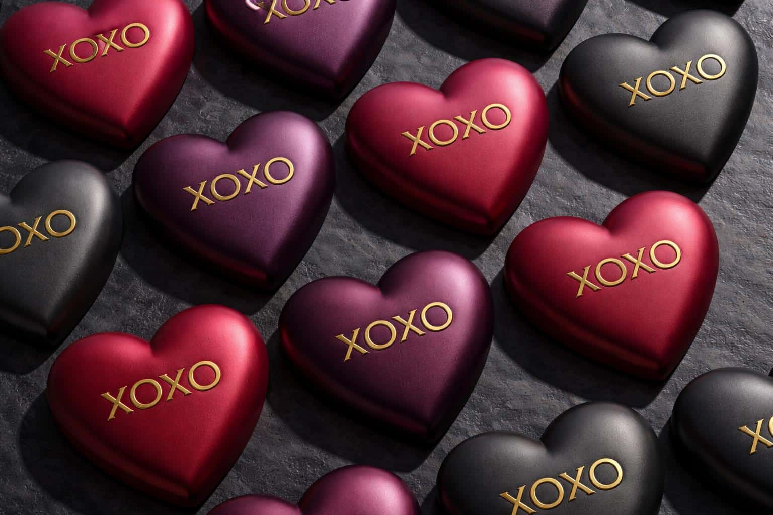 Wide 3:2 render of satin-matte Lux candy hearts in ruby, plum, and charcoal with debossed XOXO gold inlay, arranged in a geometric grid on dark stone.