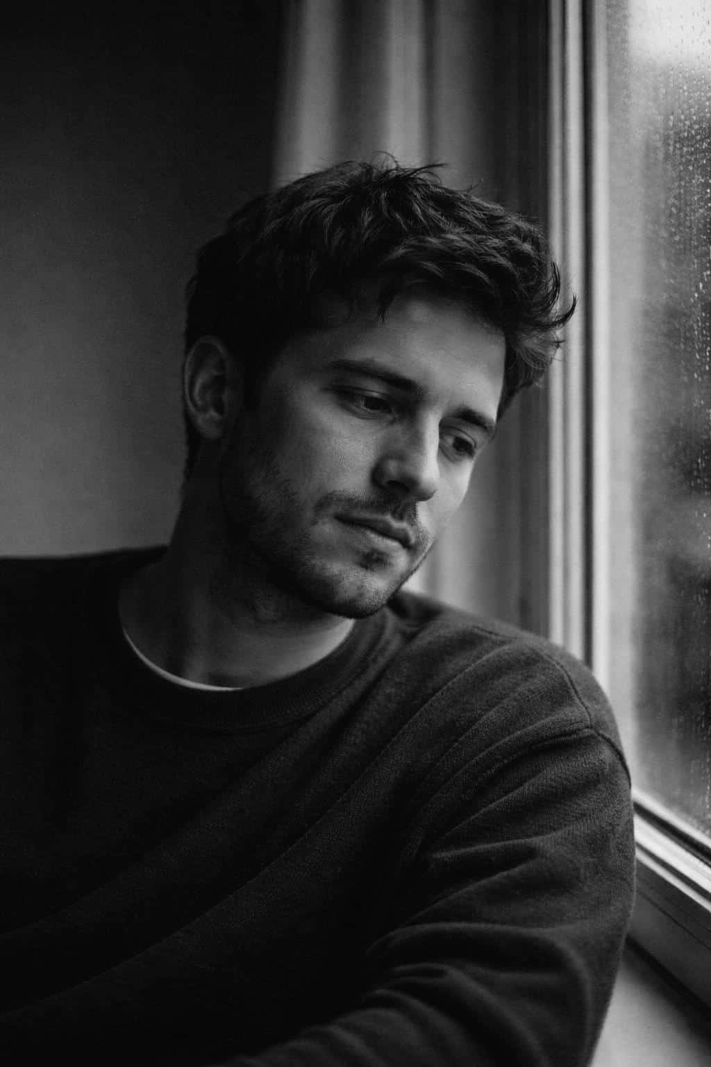 Cinematic 2:3 vertical editorial portrait of a man in his early 30s, chest-up by a rainy window with a calm, reflective expression and classic Kodak Tri‑X film grain.