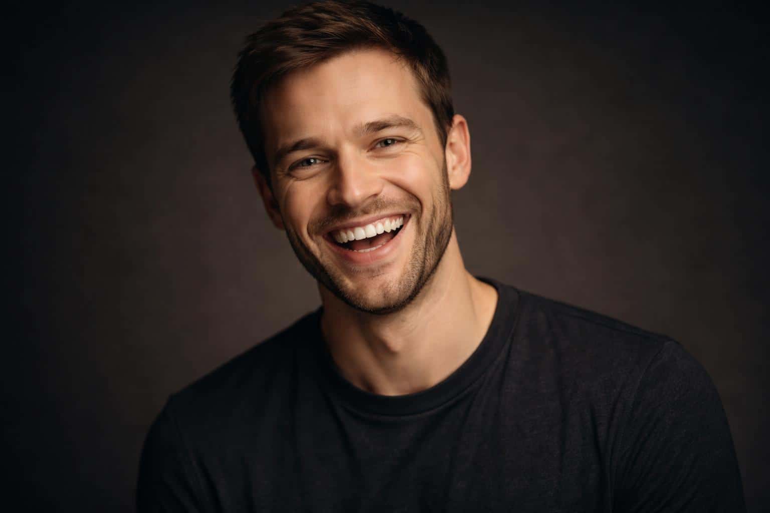 Wide 3:2 cinematic editorial studio headshot of a white man in his early 30s, tight upper-chest crop, laughing with a big teeth-showing smile and crow’s-feet, charcoal backdrop.