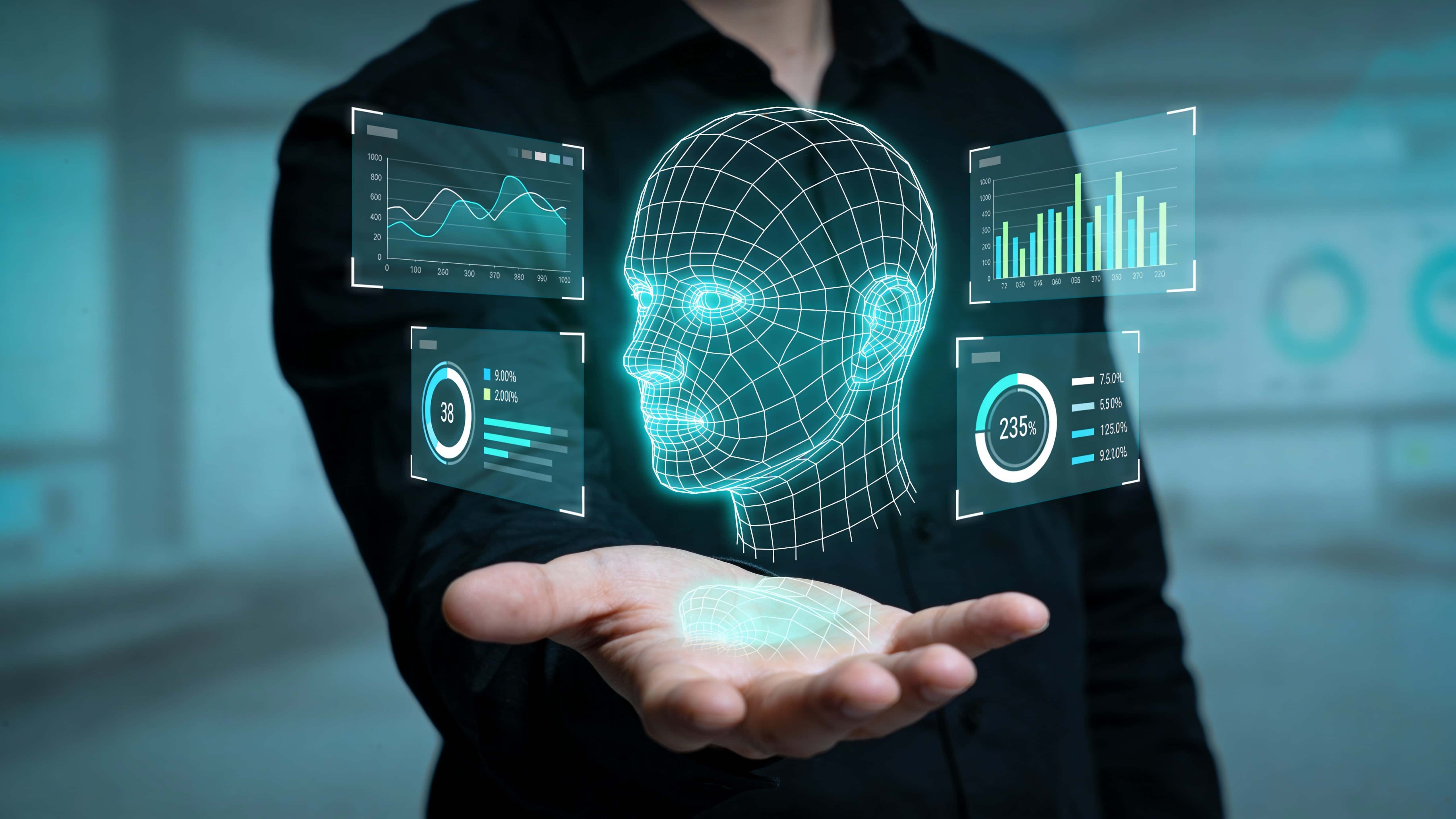 A man in a dark minimal environment holds an open hand interacting with a floating wireframe human head glowing cyan and teal, surrounded by transparent data panels.