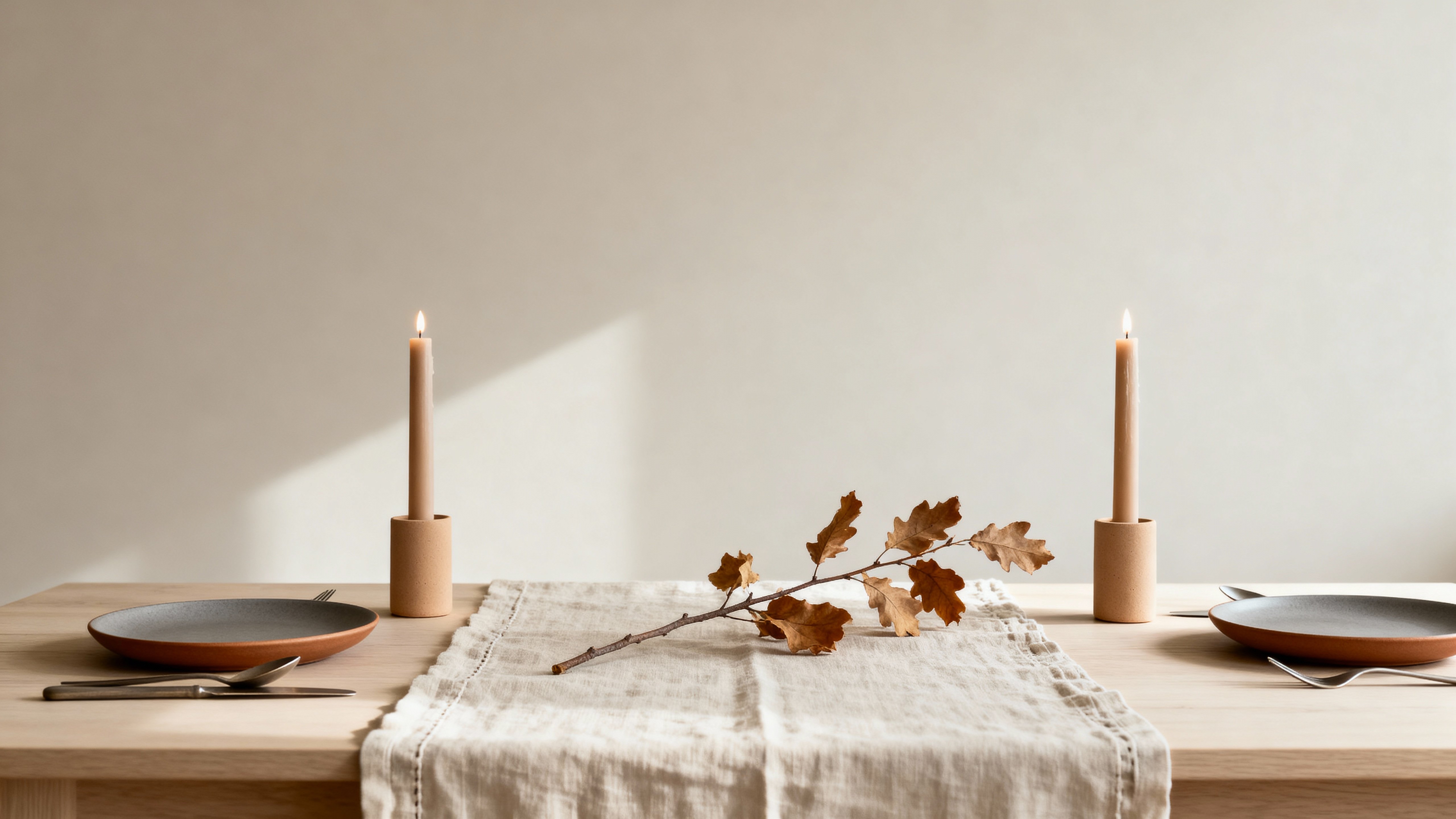 A minimal tablescape for a small seasonal editorial: undyed linen and matte warm-gray stoneware, sand candles and dried-branch centerpiece on a pale-wood table with creamy, low-contrast light.