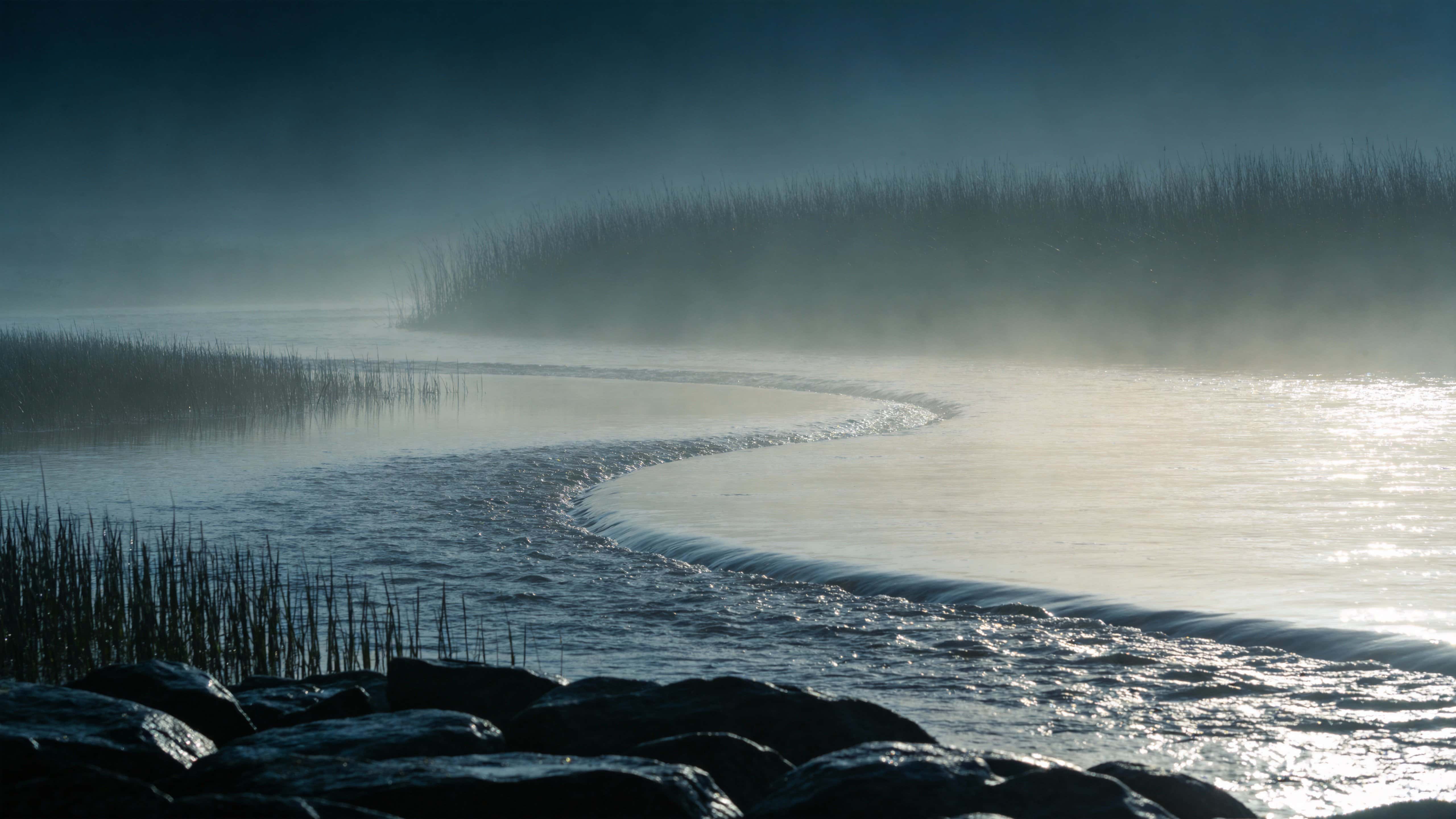 A 16:9 landscape photograph of a mist-shrouded river at dawn, captured with soft diffused light and calm reflections for a serene wallpaper-ready composition.