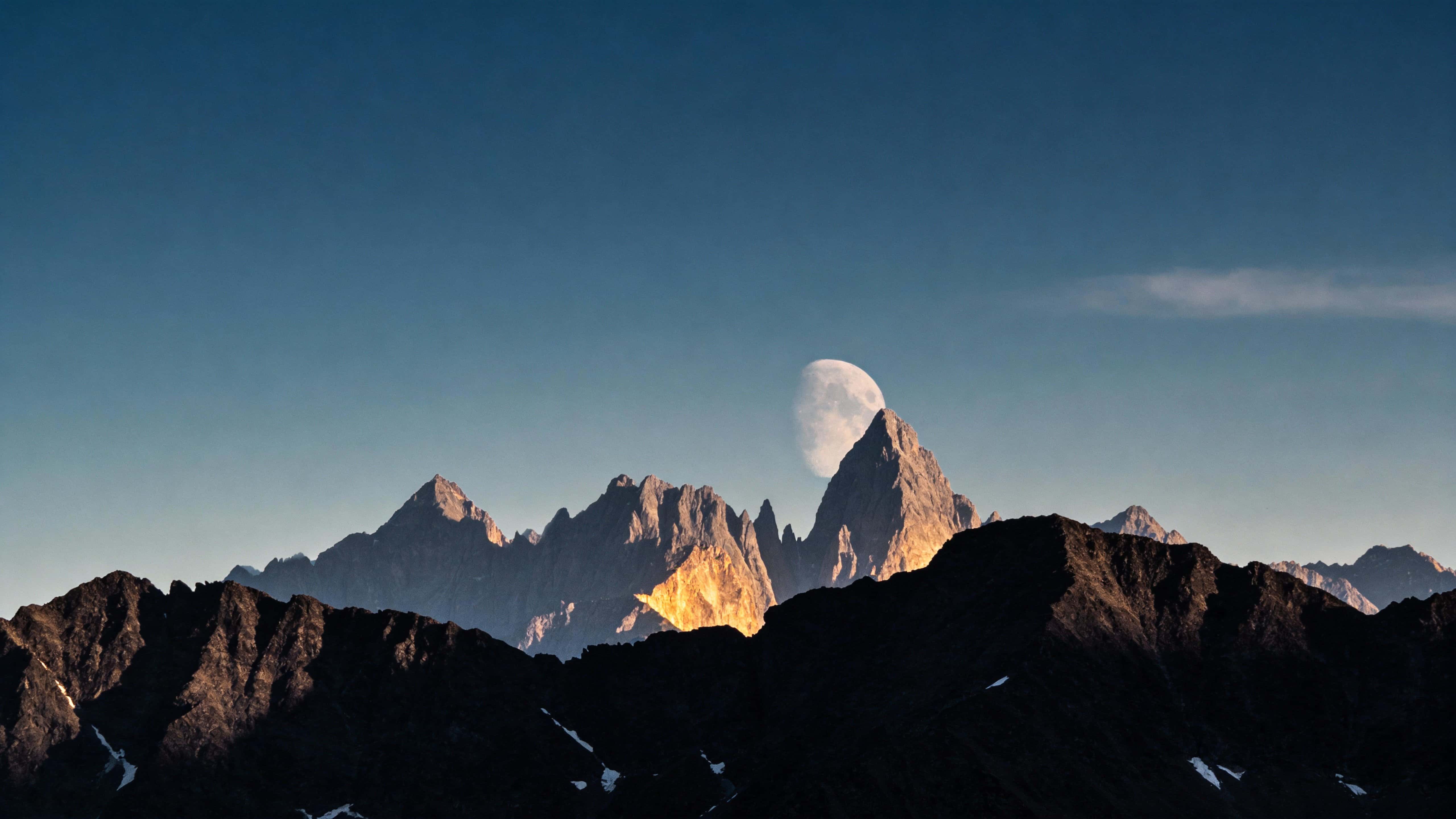 Moon rising behind rugged mountain peaks, framed as a widescreen landscape photograph for a 16:9 wallpaper that emphasizes dramatic backlight and a serene night mood.